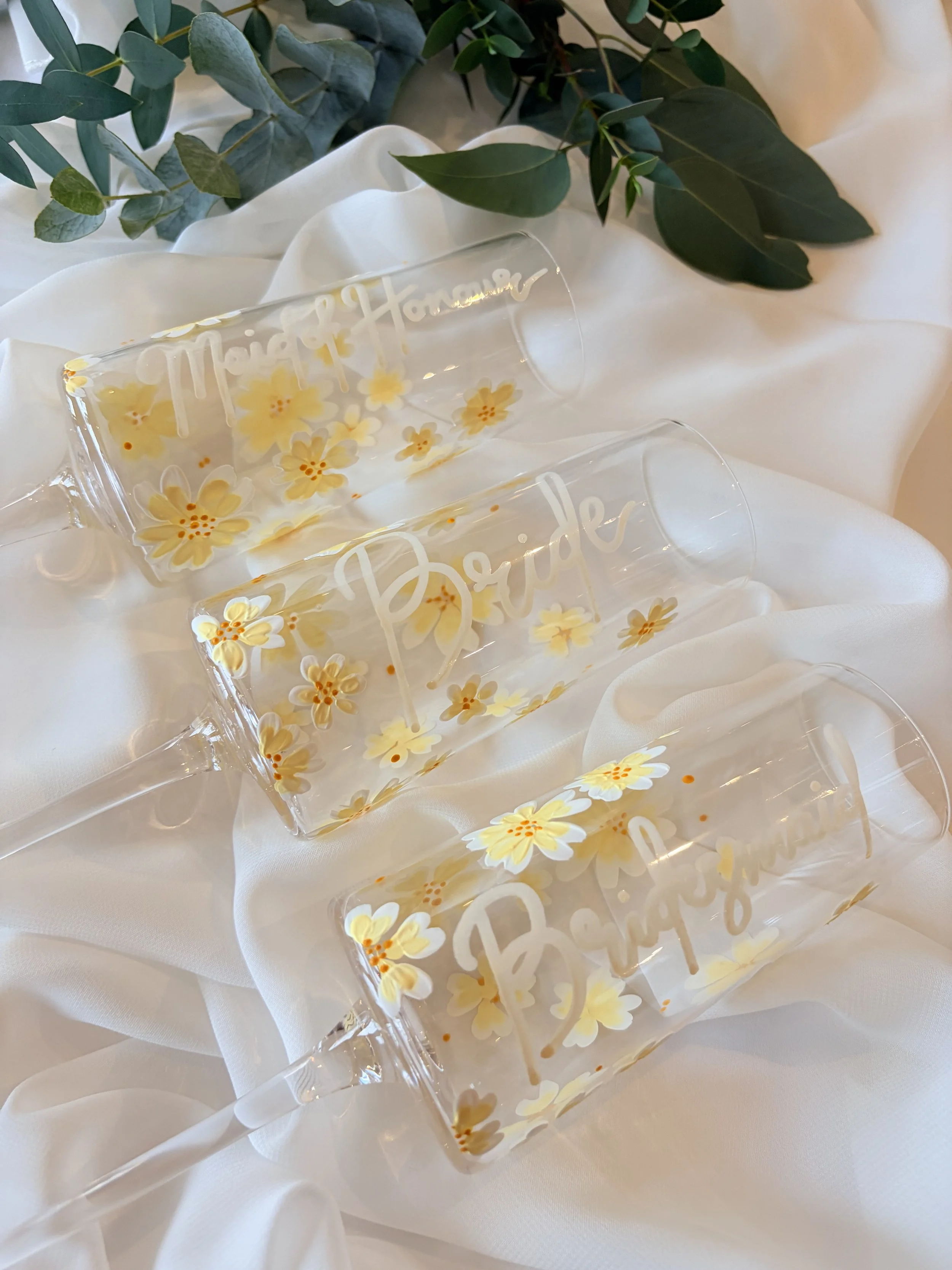 Three champagne flutes decorated with yellow and white flowers and the words "Thank You" and "Brides" written on them, resting on white fabric with a leafy green branch in the background.