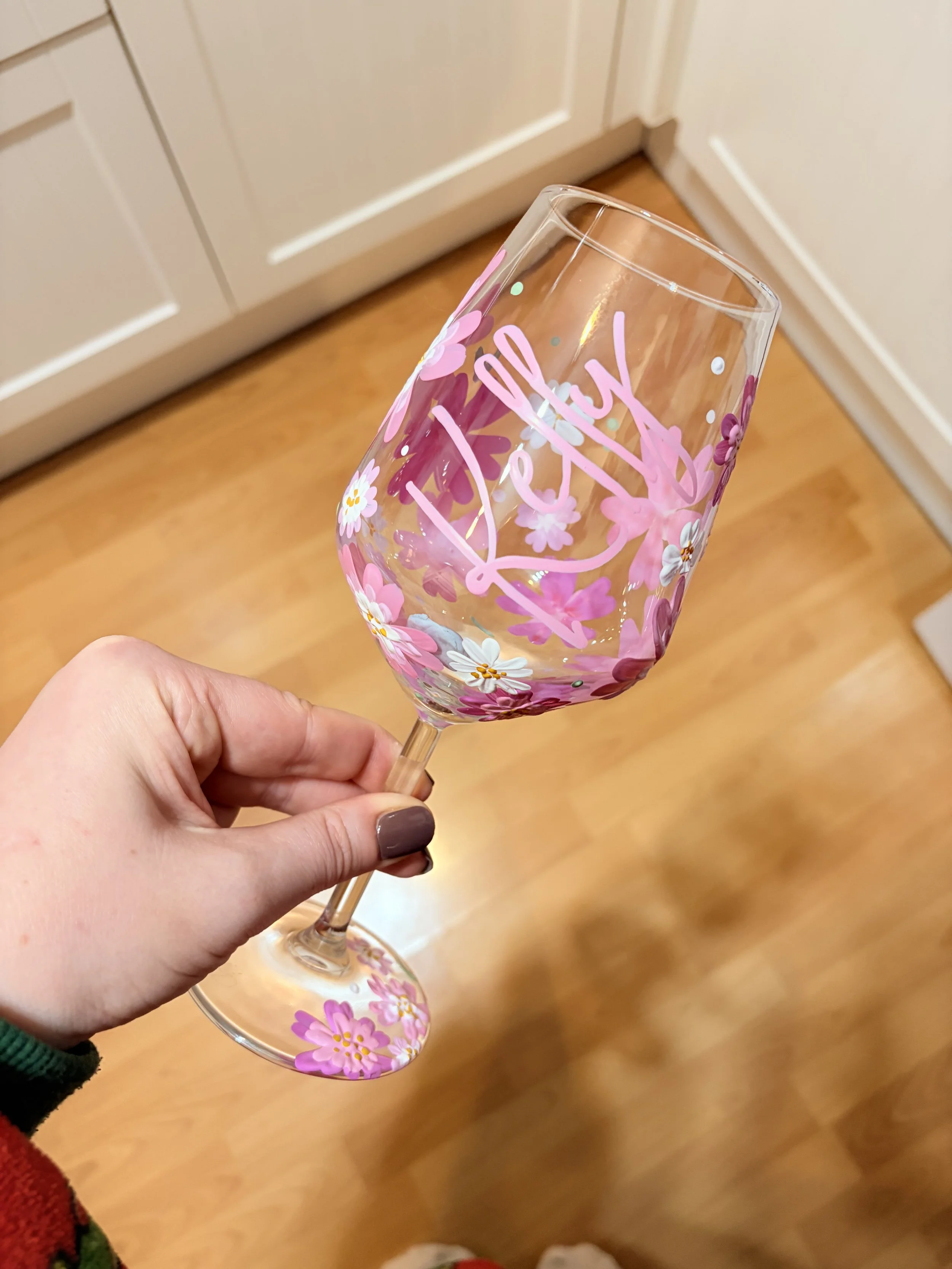 Hand holding a decorated wine glass with floral patterns and the words 'Thank You' written on it, in a room with wooden flooring and white cabinets.