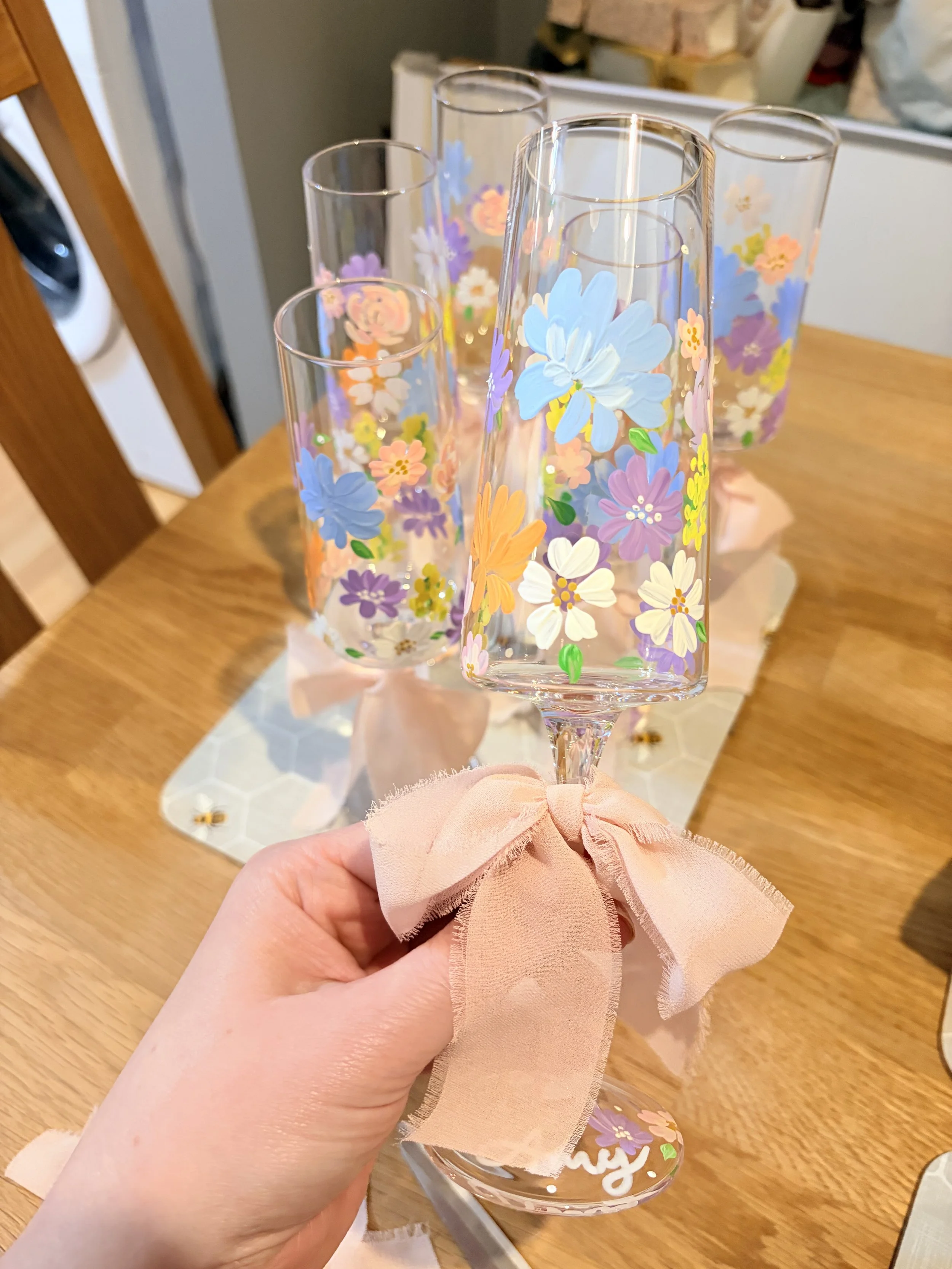 Hand holding a decorated champagne flute with colorful floral designs, tied with a peach-colored fabric ribbon, on a wooden table with a floral centerpiece.