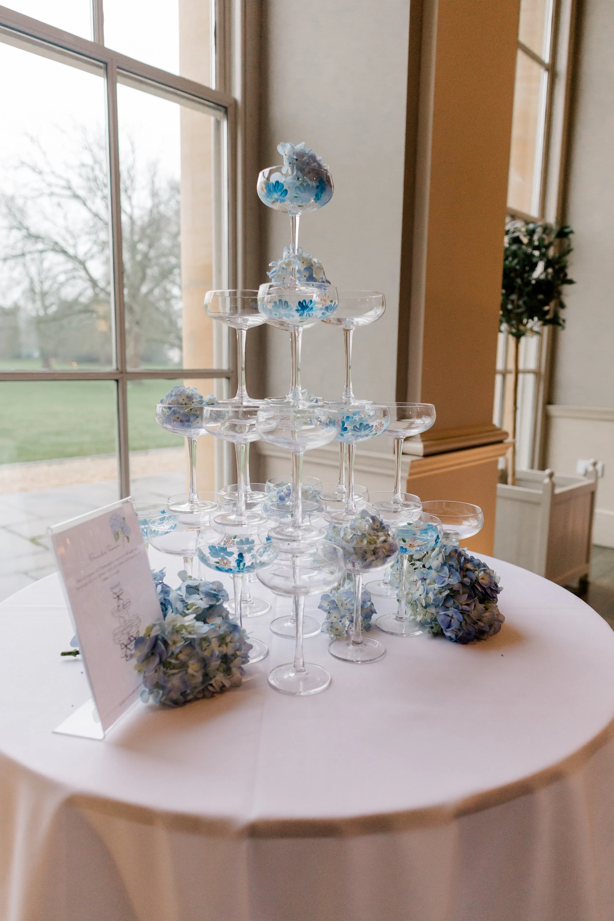 A tiered display of empty champagne glasses with blue and white hydrangea flowers around a white tablecloth, set near large windows with a view of a grassy outdoor area.
