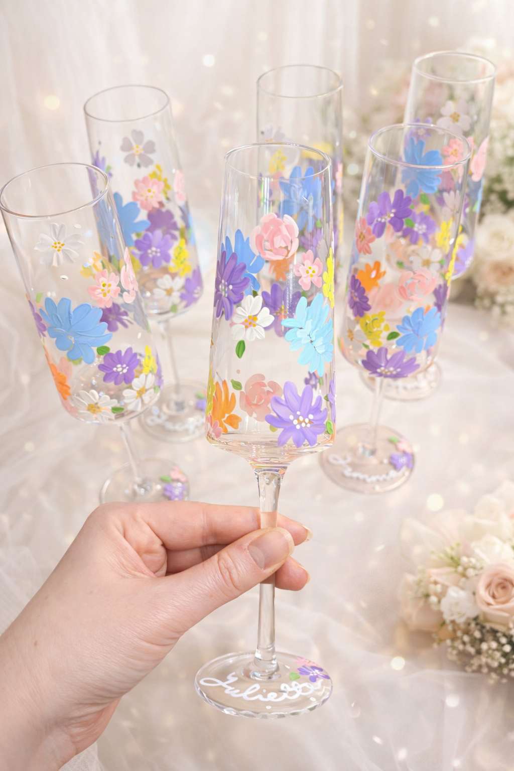 Hand holding a decorated champagne flute with floral patterns, set among other similar glasses on a table with a soft light background.