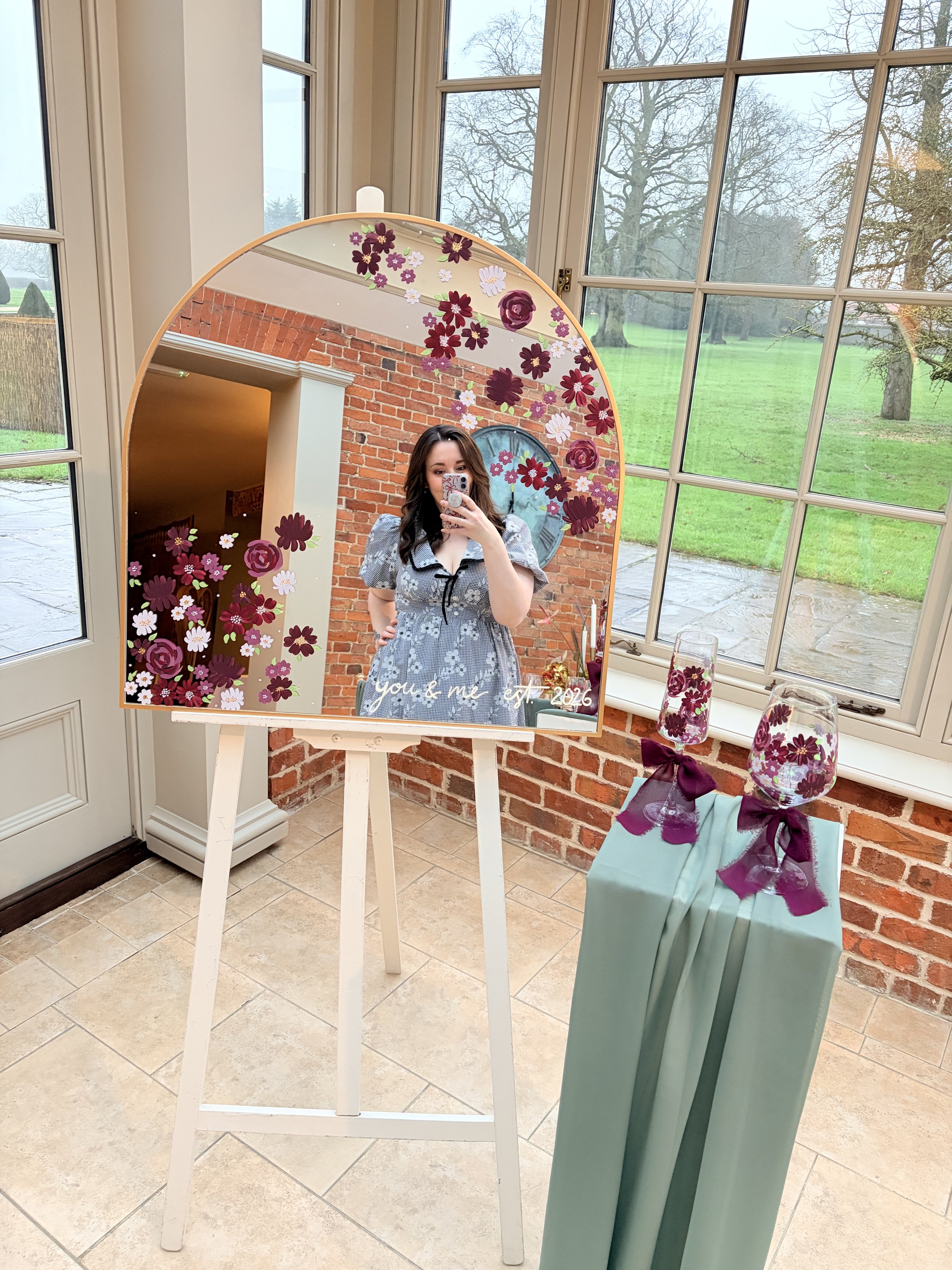 A woman taking a selfie in a mirror decorated with floral stickers, with a brick wall and large windows revealing an outdoor green landscape in the background.