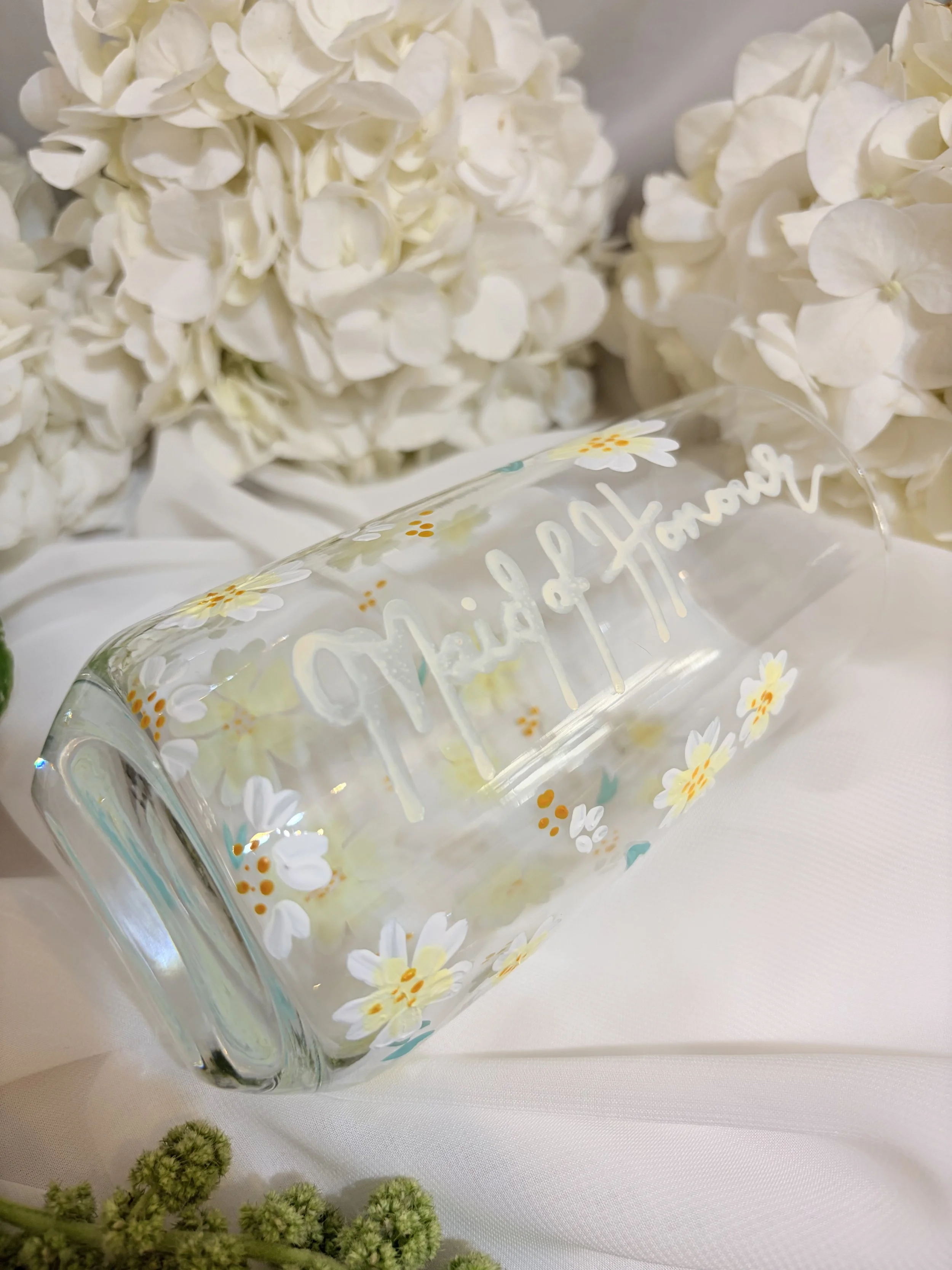 A glass with the words 'Happy Honey' written on it, decorated with daisies, placed in front of white hydrangea flowers on a white surface.