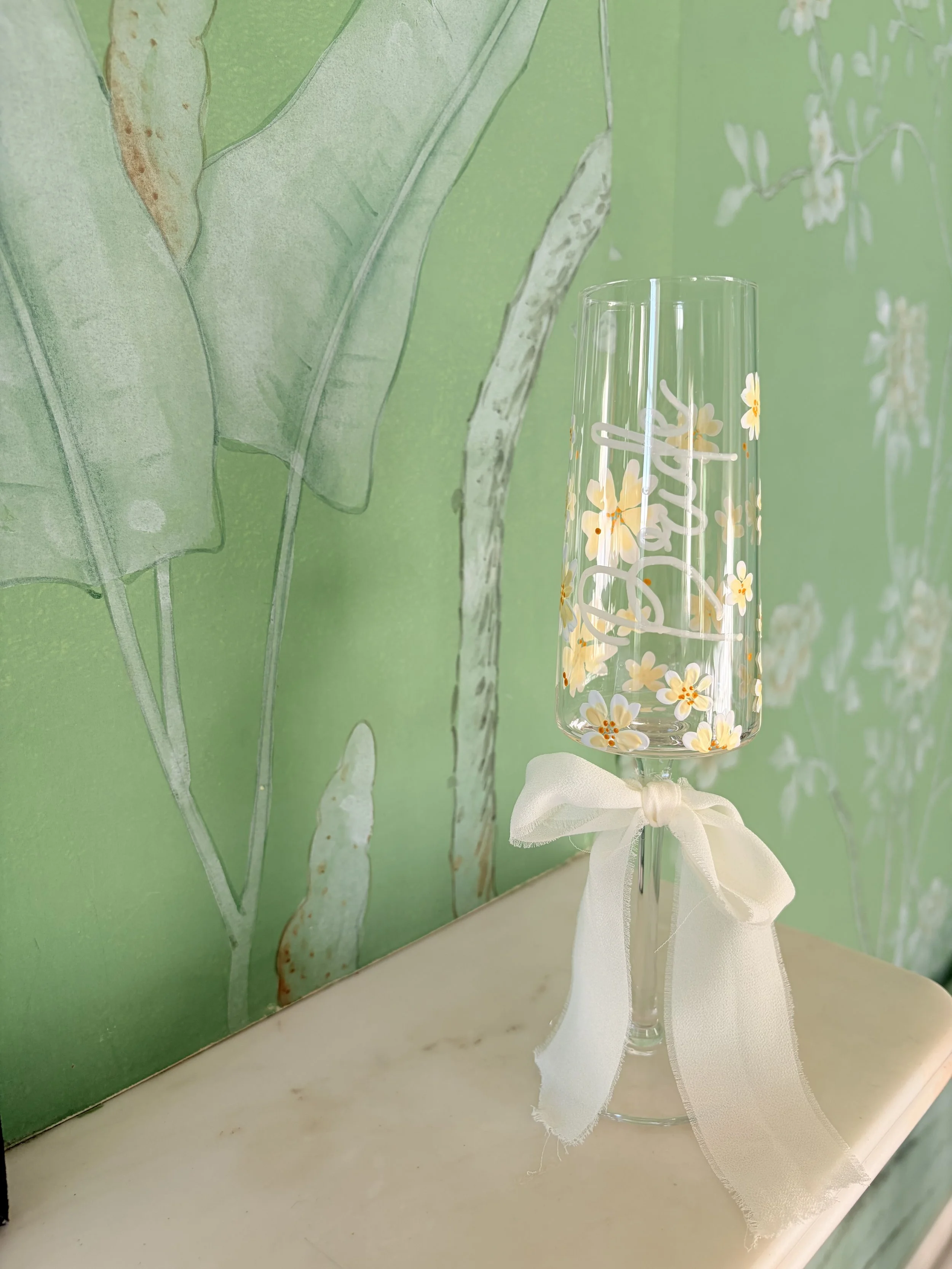 A tall glass vase with a white bow around its neck, decorated with white and yellow flowers, sitting on a white surface with a green painted leafy plant design in the background.