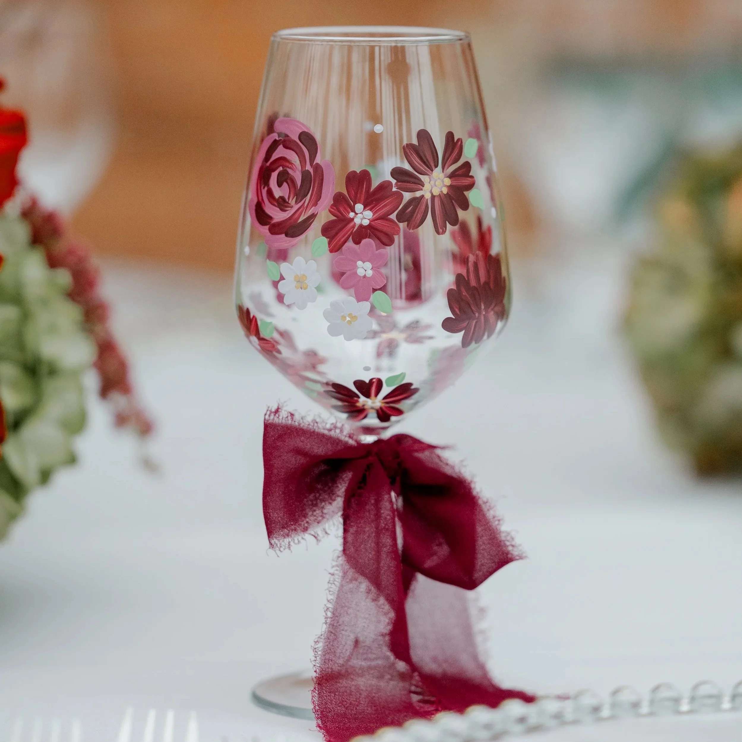 A clear wine glass decorated with pink and red floral patterns, tied with a red ribbon at the base, placed on a white surface with blurred floral arrangements in the background.