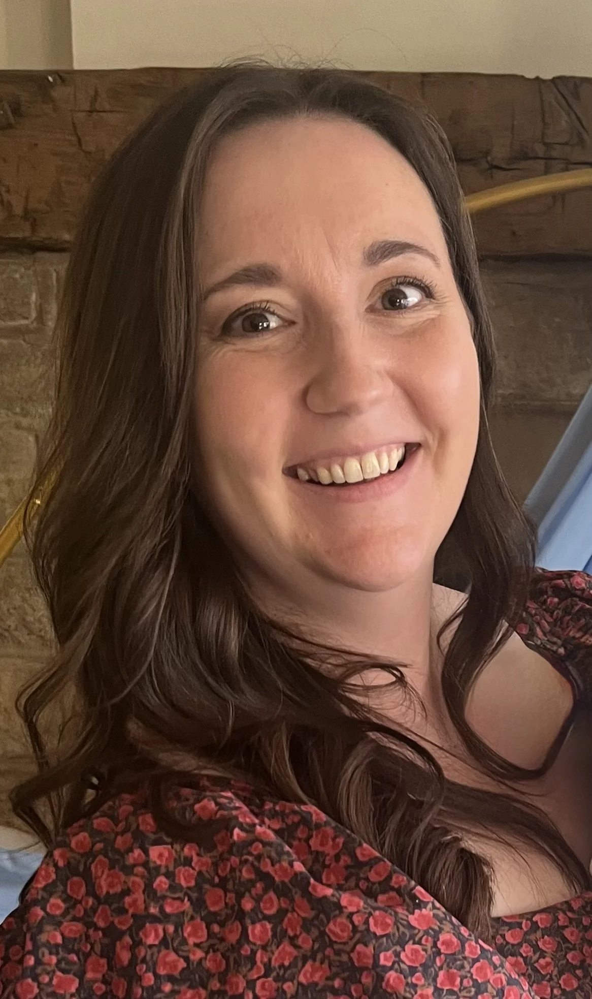 A woman with long, wavy brown hair smiling at the camera, wearing a red and black floral top, with a wooden and brick background.