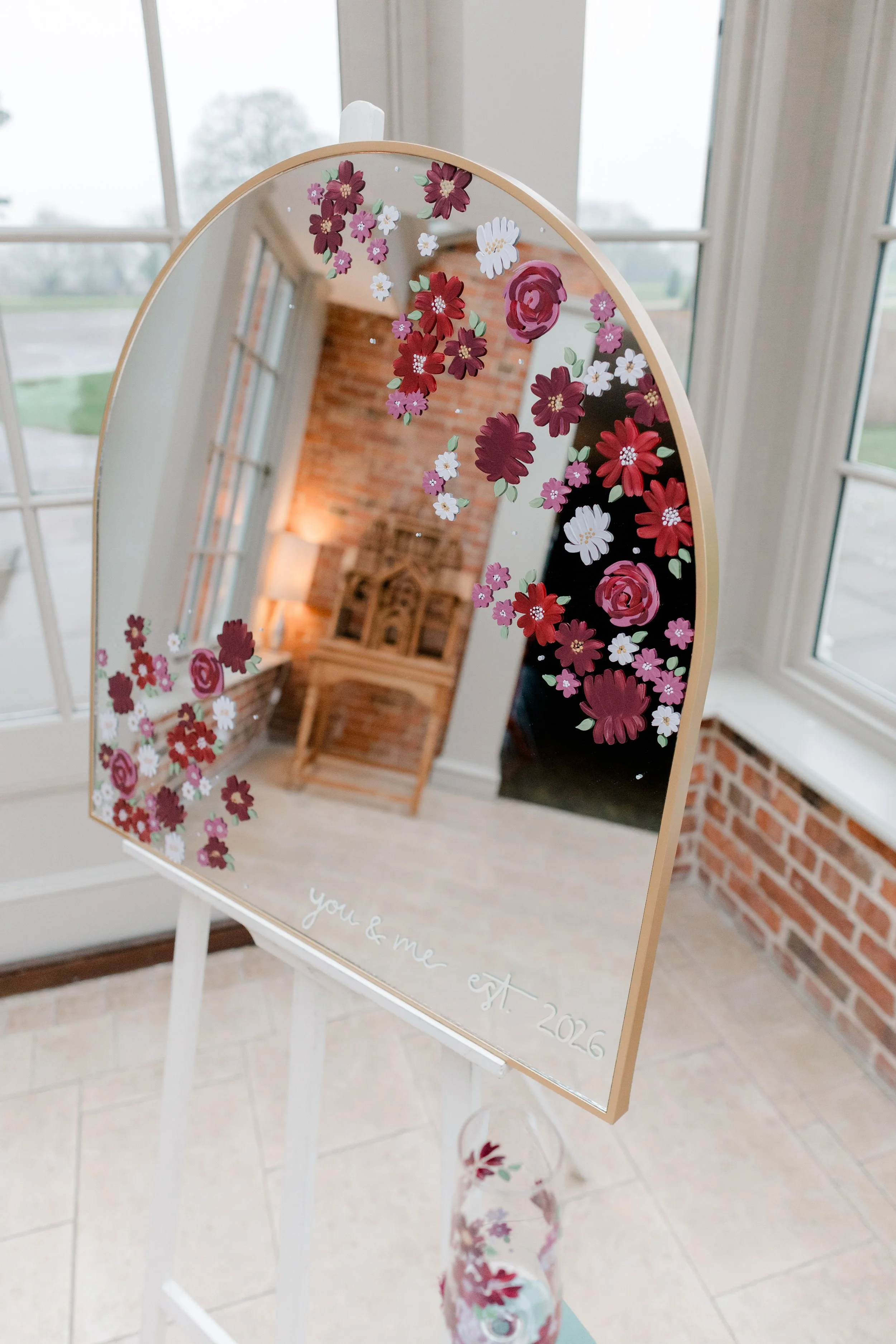 Decorative mirror with flower designs and the text "you & me est. 2016" reflected in a room with brick walls, a wooden chair, and large windows showing an outdoor scene.