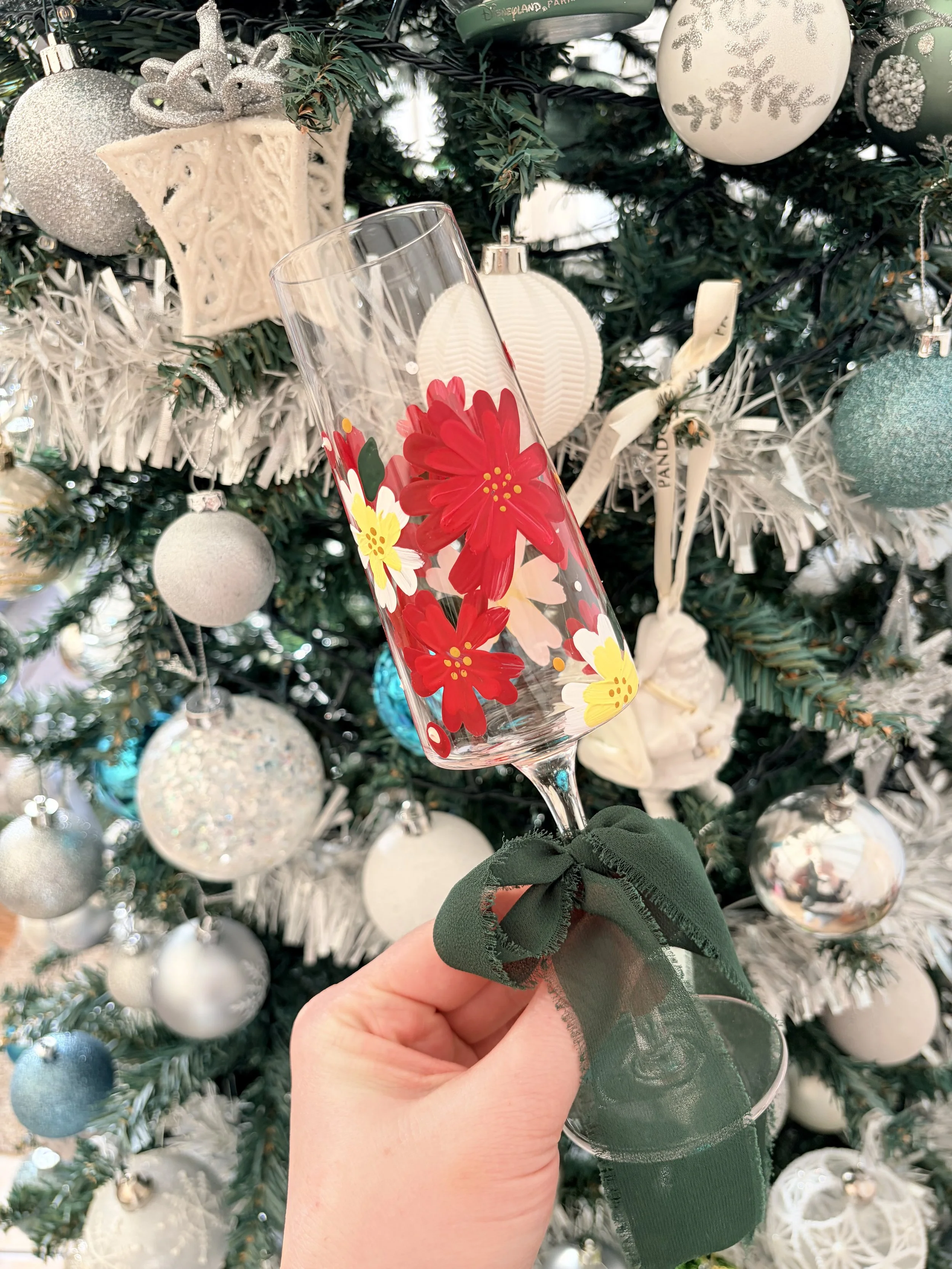 Person holding a decorated champagne flute with red and yellow floral designs, green ribbon around the stem, in front of a Christmas tree with white, silver, and teal ornaments.