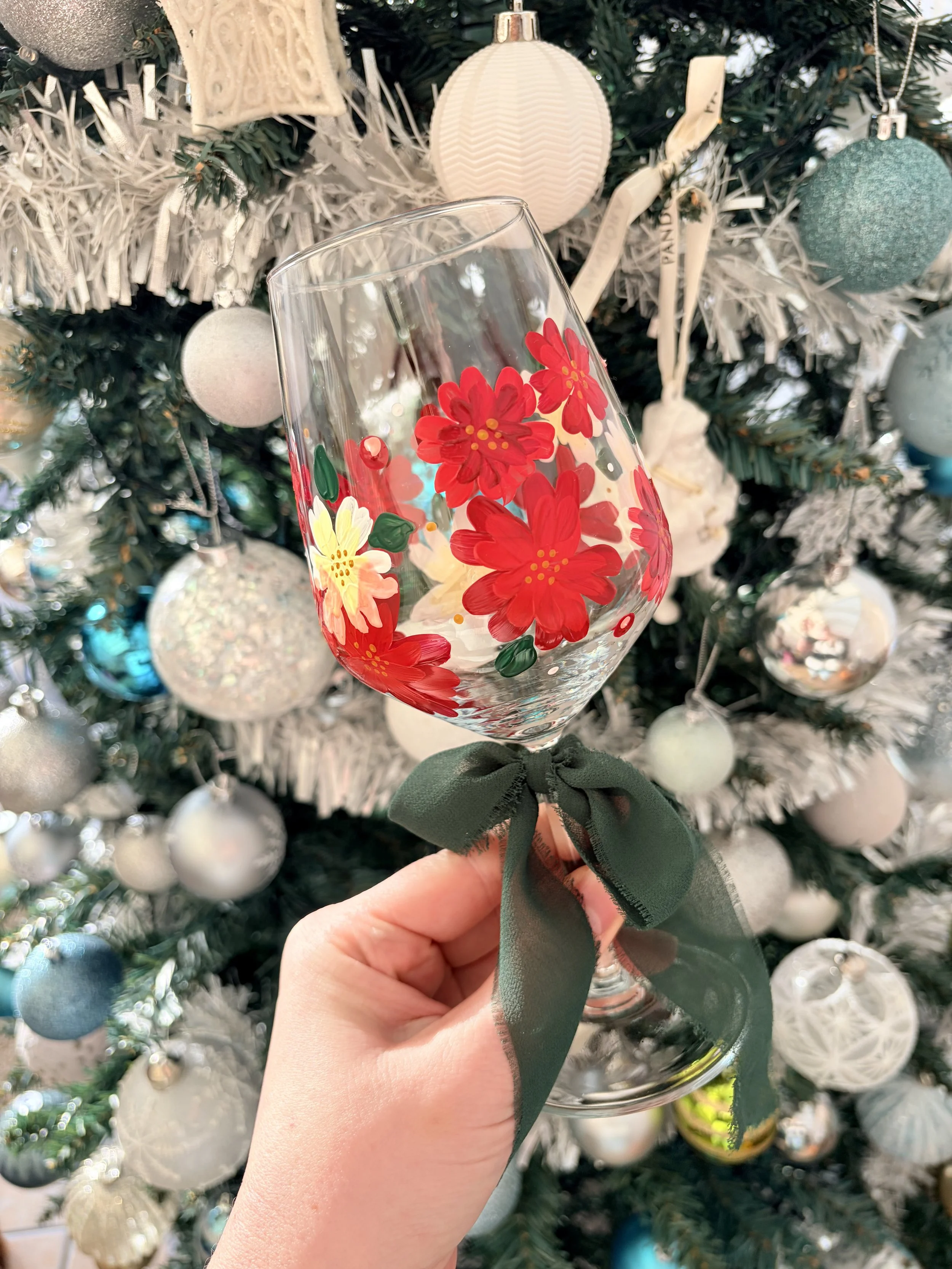 Hand holding a glass decorated with red and yellow flowers, tied with a dark green ribbon, in front of a white decorated Christmas tree with ornaments.