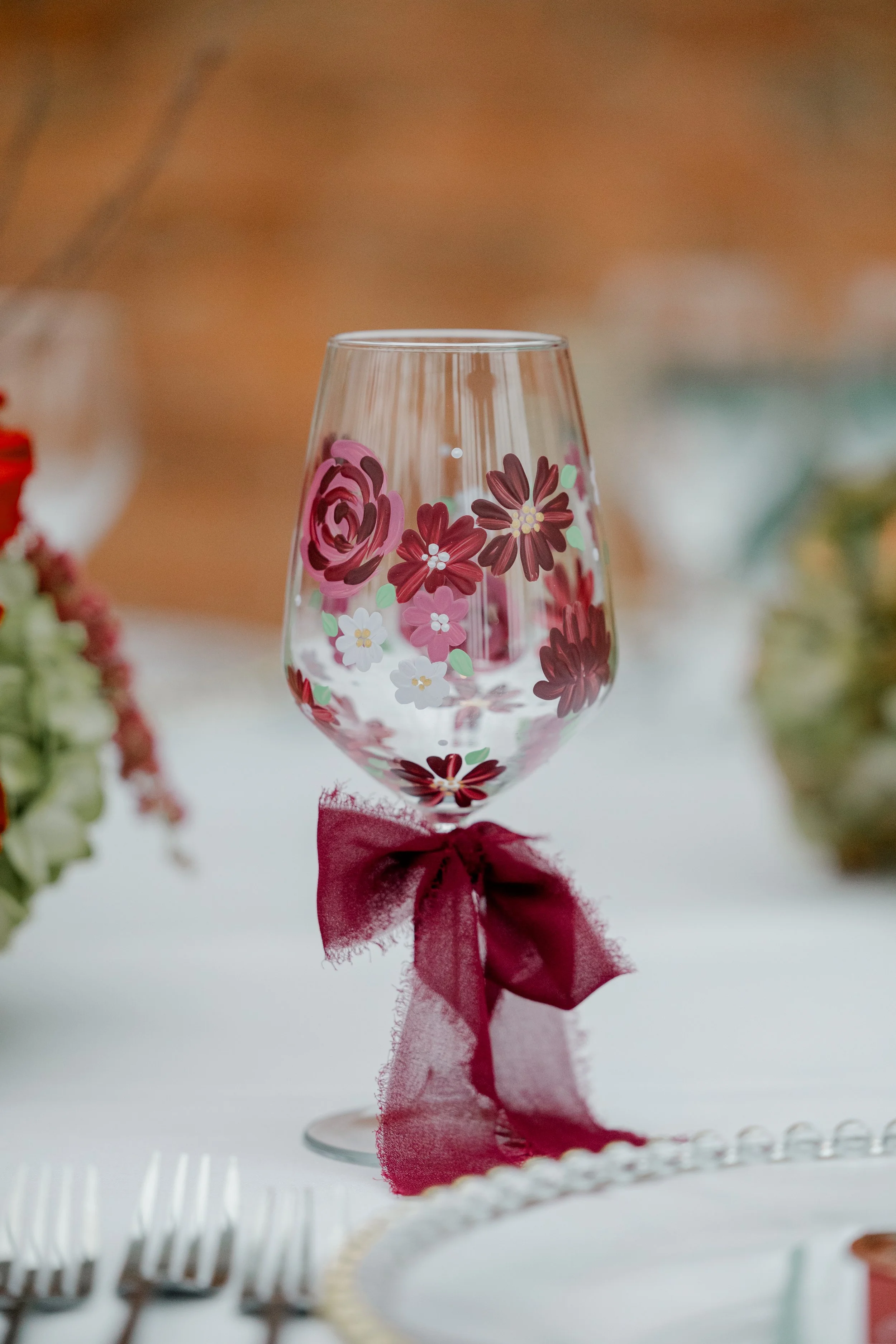 A clear wine glass decorated with pink and red floral patterns, tied with a maroon ribbon at the stem. The glass is set on a table with blurred colorful flowers on either side.