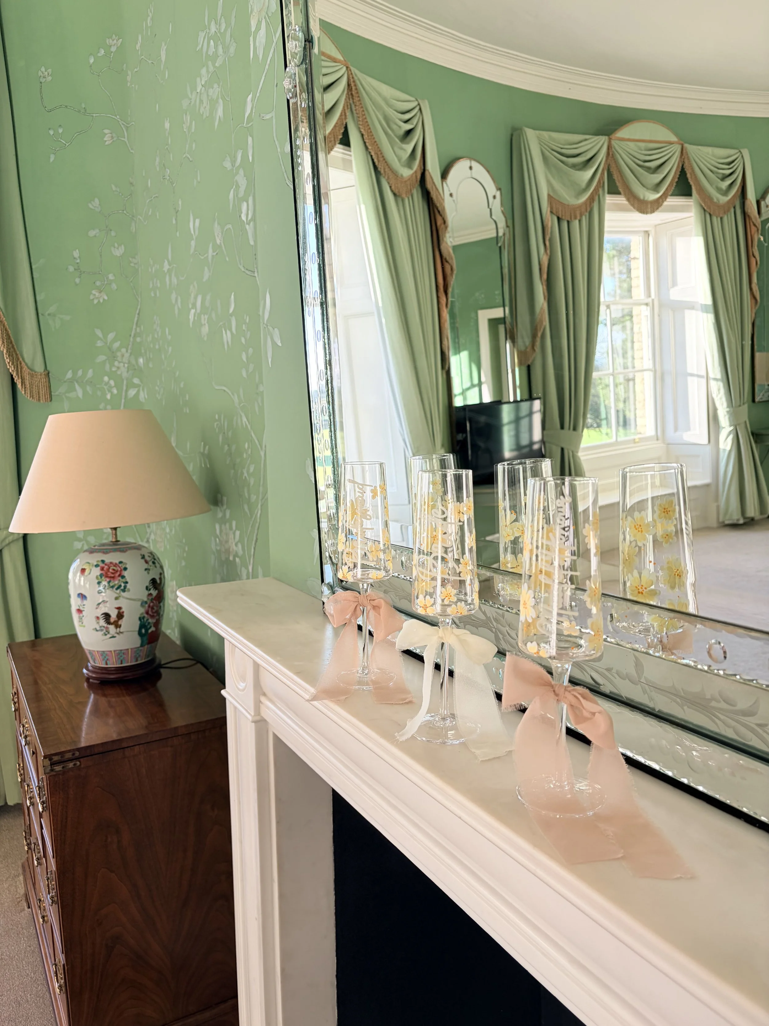 Decorative room with window and green curtains, reflecting in a large mirror; small champagne glasses with daisy pattern and pink bows on the ledge, and a floral lamp on a wooden side table.