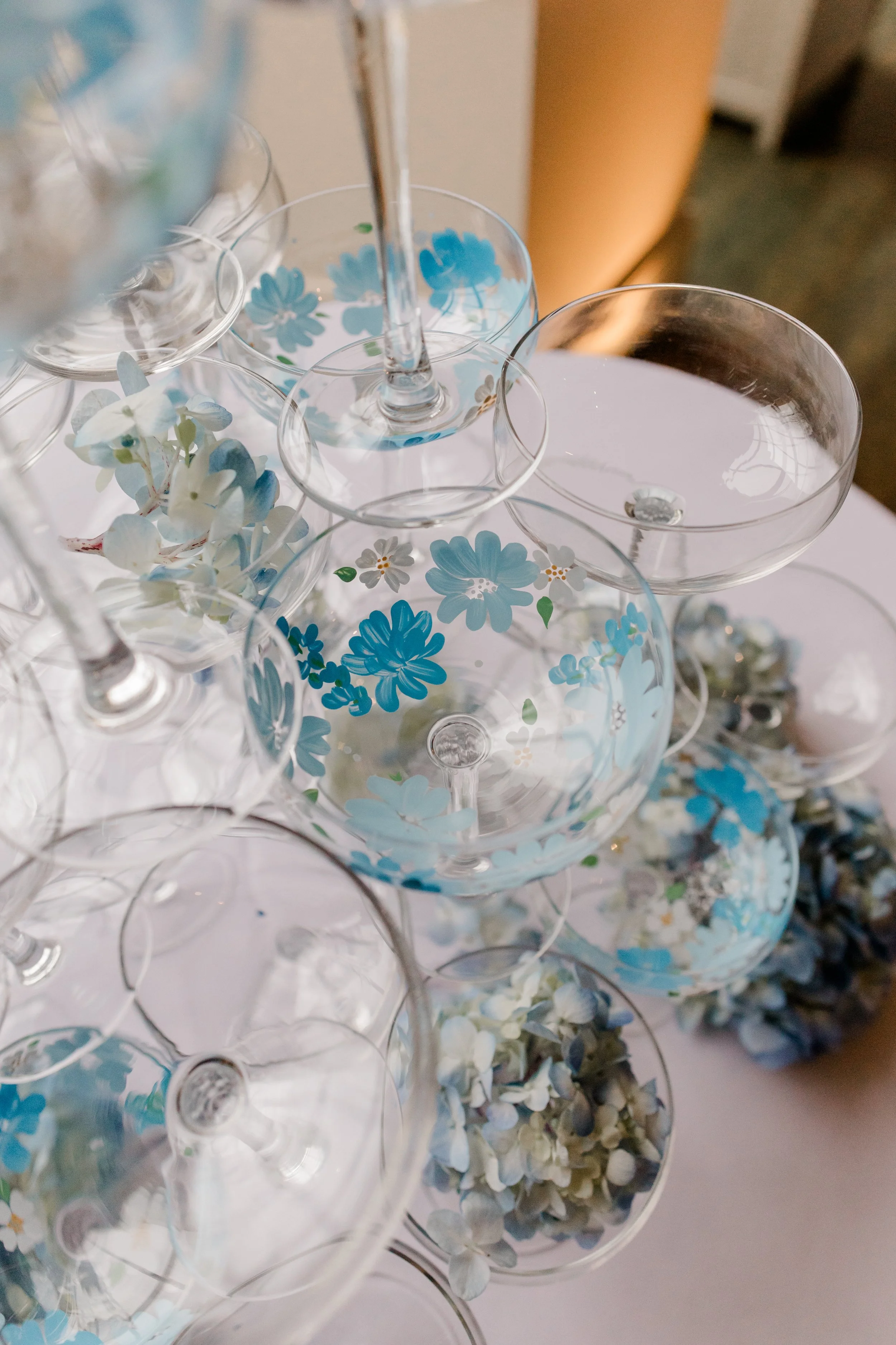Pile of empty wine glasses with blue floral patterns and white and light blue flowers scattered around on a white surface.