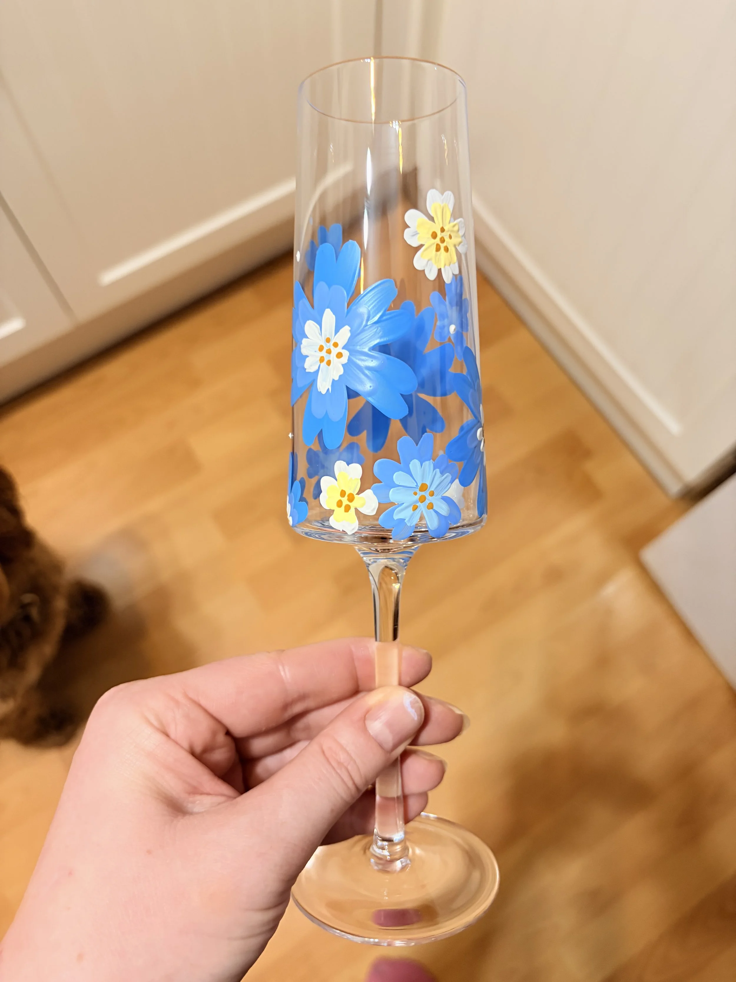 A hand holding a champagne flute with blue and white flowers painted on it, in a room with wooden flooring and a white door in the background.