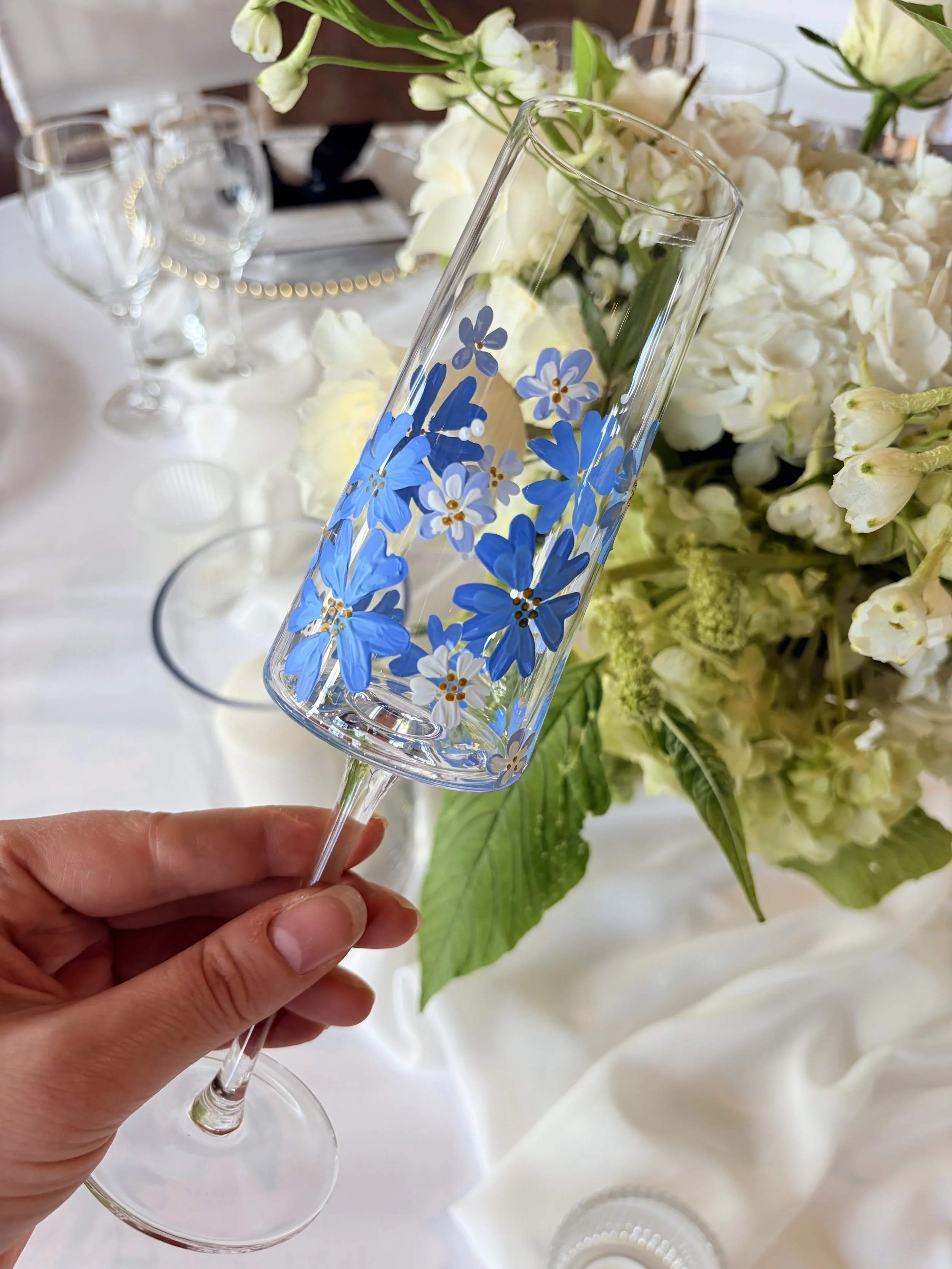 A hand holding a champagne flute decorated with blue and white floral patterns, with a floral arrangement of white flowers in the background