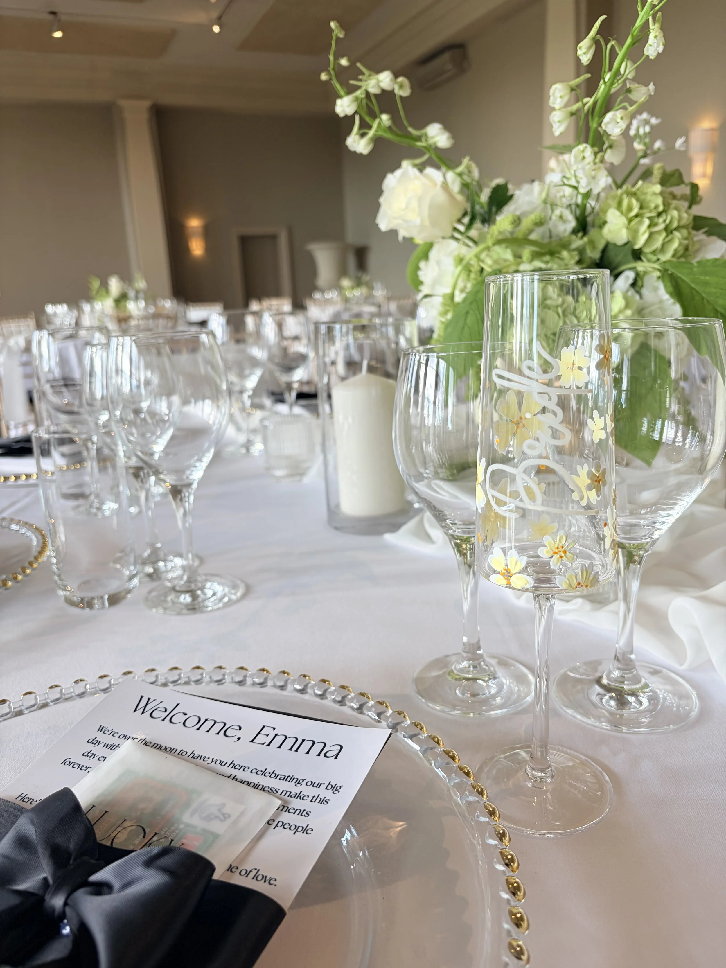 Elegant dining table set with glassware, a floral centerpiece with white and green flowers, candles, and a welcome card for Emma at the table.