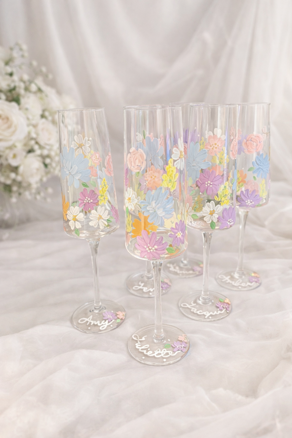 Four champagne flutes decorated with colorful floral designs and personalized with names written on the bases, arranged on a white fabric surface with a bouquet of white flowers in the background.
