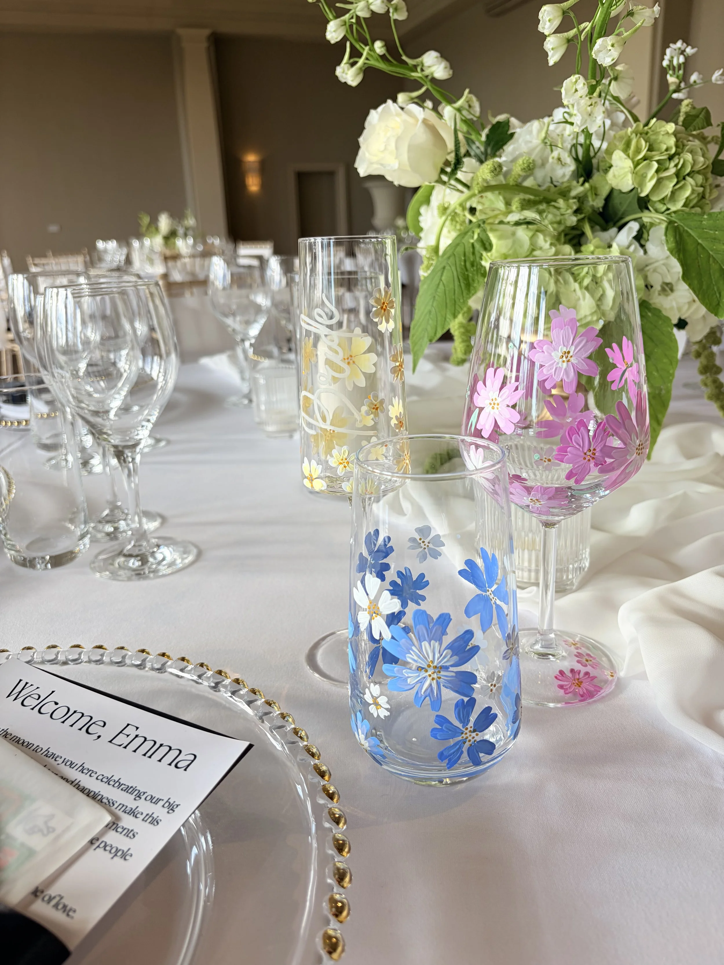 Decorative glasses with colorful floral patterns on a wedding or event table, with a floral centerpiece in the background and a welcome card.