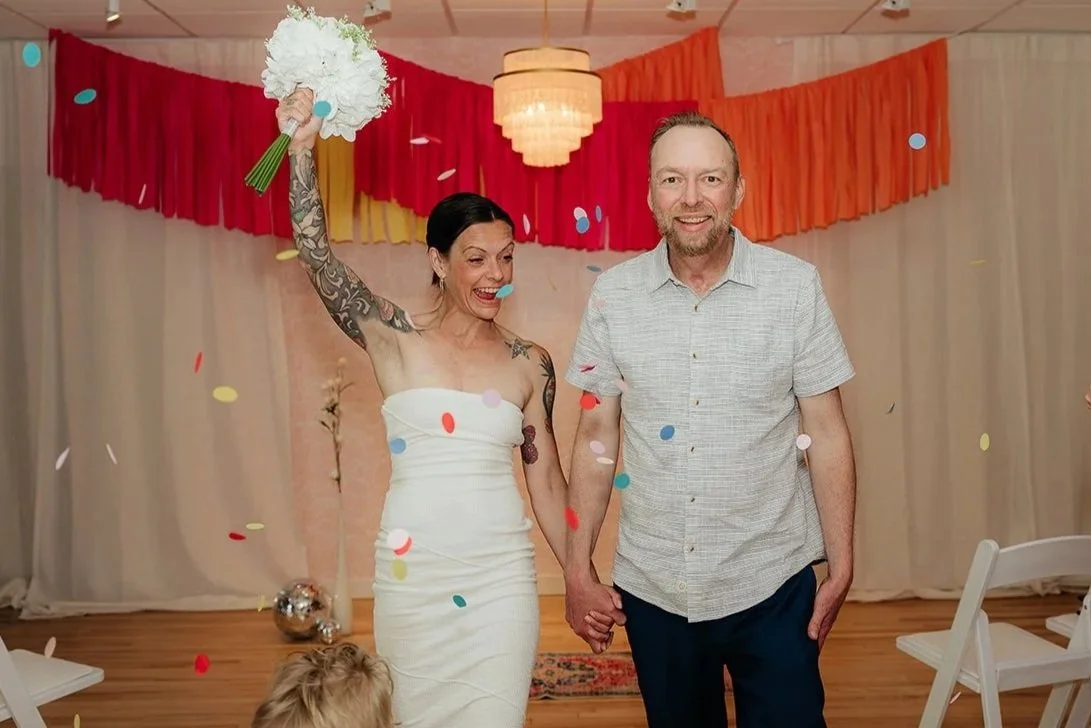 A woman in a white dress holding a bouquet with her right hand raised, holding hands with a man in a light-colored shirt, both smiling at a celebration with colorful confetti and a decorated background.
