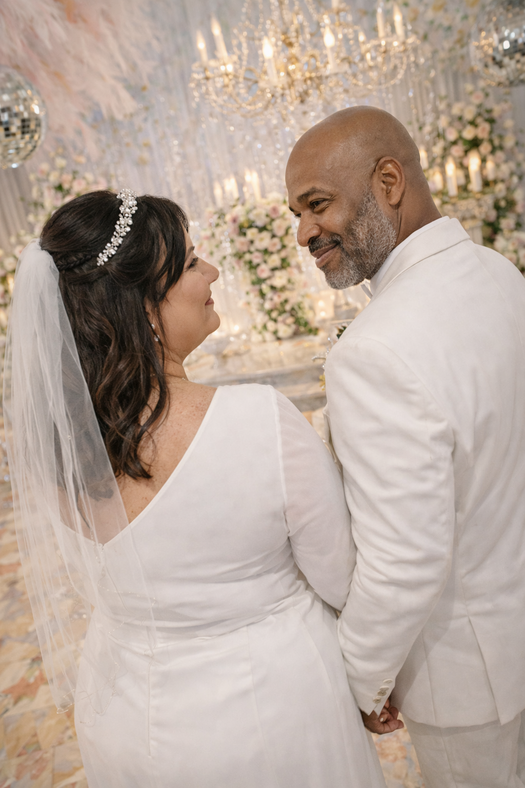 A bride and groom holding hands, gazing into each other's eyes during their wedding ceremony in a decorated venue with chandeliers and floral arrangements.