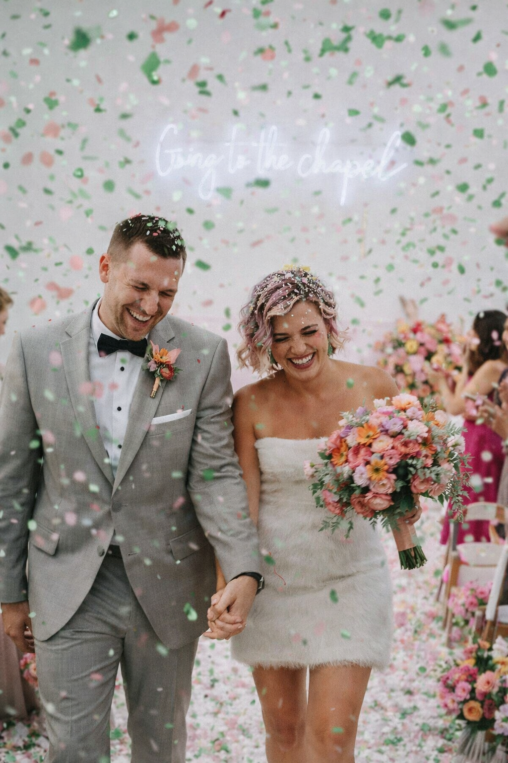 A newlywed couple, a man in a gray suit with a black bow tie and a woman in a strapless white dress holding a bouquet, walk hand in hand through falling pink, green, and white confetti at their wedding reception with a pink floral backdrop and guests in the background.