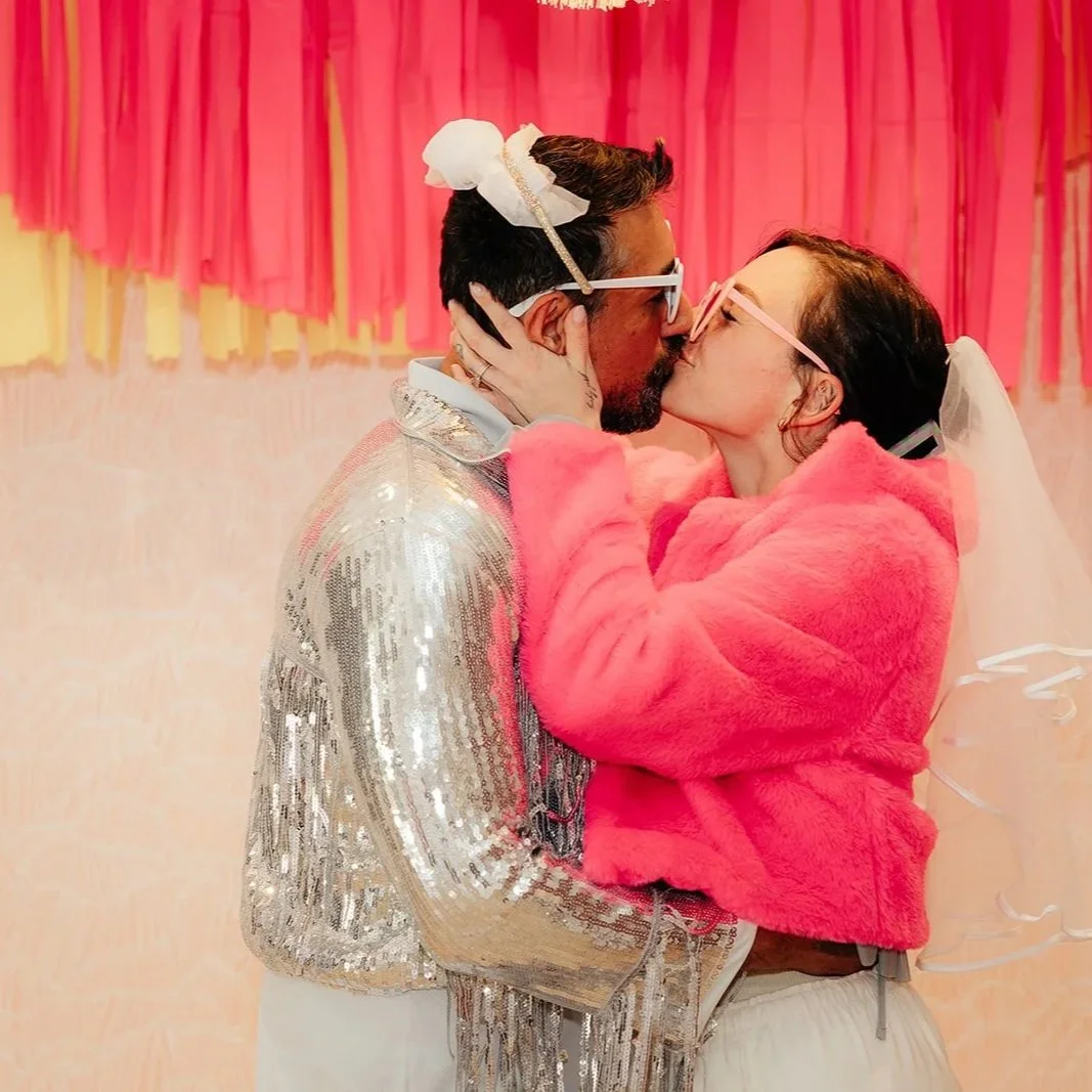 A couple kissing at their wedding reception, dressed in colorful attire, with decorations in the background.