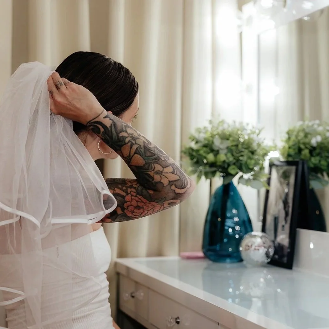 A woman with tattoos on her arms, wearing a veil, appears to be getting ready for her wedding in front of a dressing table with flowers and picture frames.