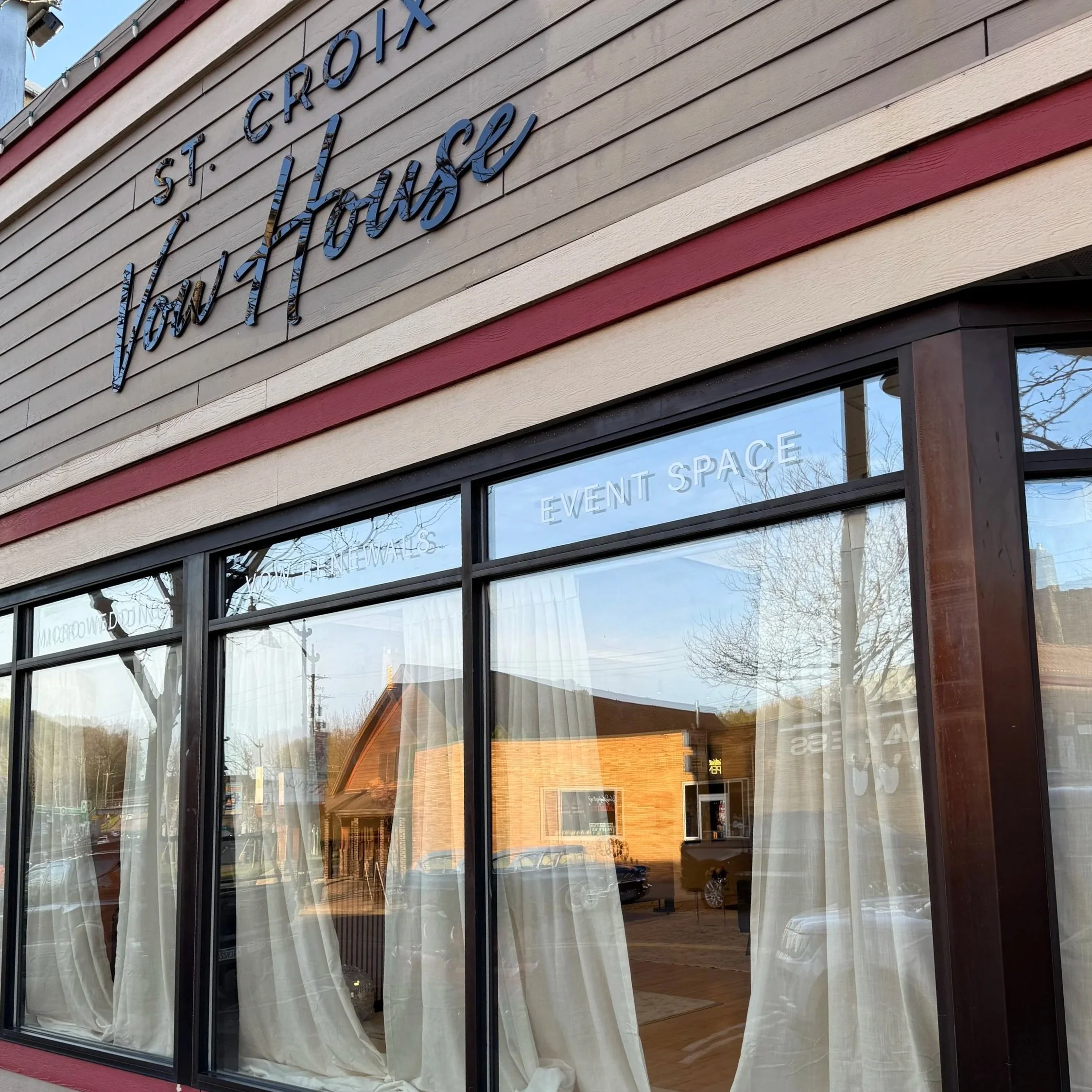 Exterior of a building with horizontal beige, tan, and red siding and large windows. The sign on the building reads 'ST. CROIX VILLAGE' in large letters. Behind the window, white curtains can be seen and the words 'EVENT SPACE' are visible.