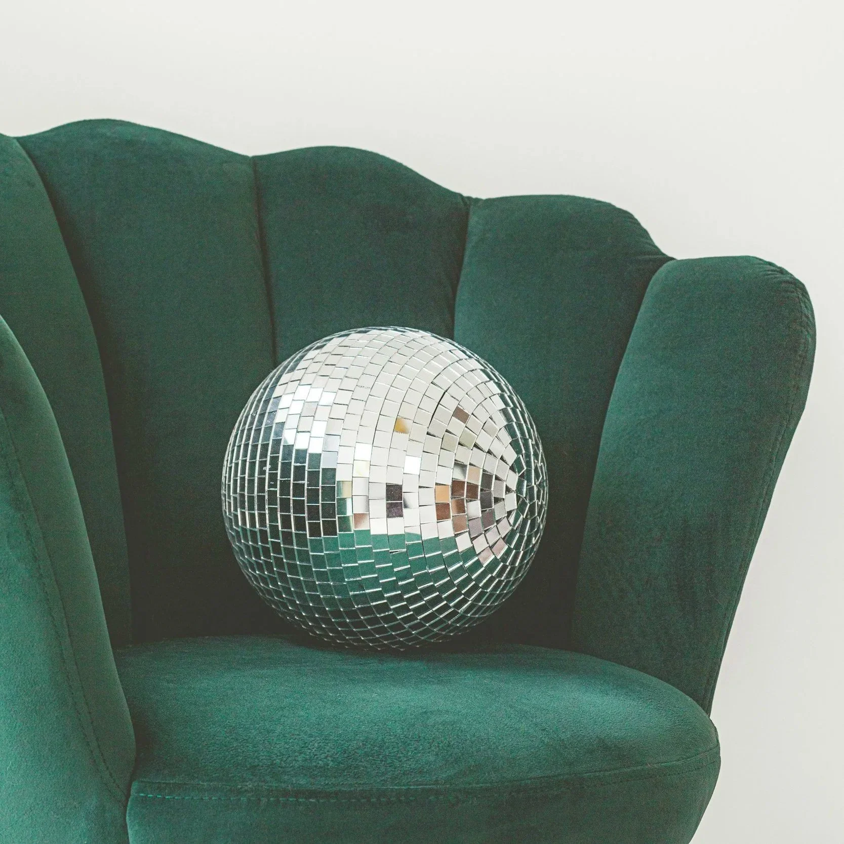 A silver disco ball resting on a dark green velvet armchair with curved backrest, against a plain white wall.