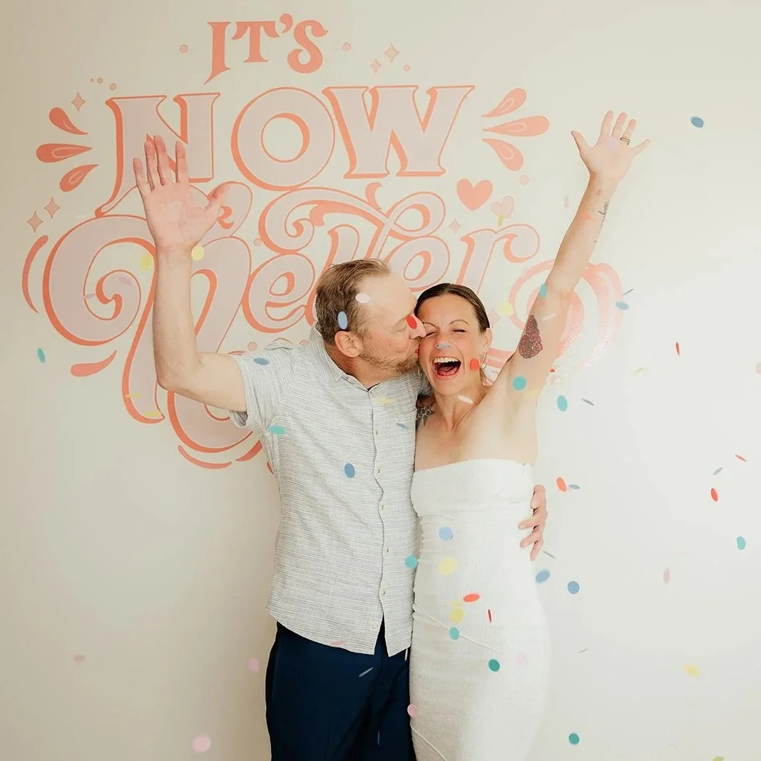 A couple celebrating with confetti, in front of a wall with 'It's now or never' written on it, smiling and embracing each other.