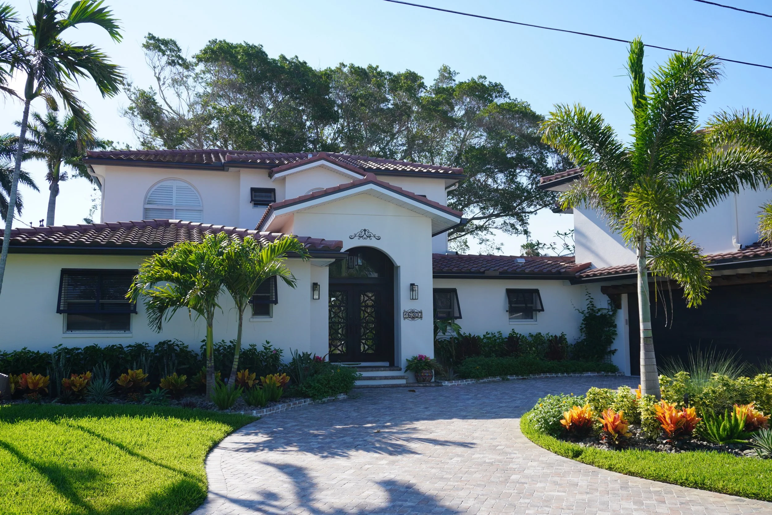Photo of a white house with red tile roof, black window shutters, and a palm tree in the front yard, under a blue sky.