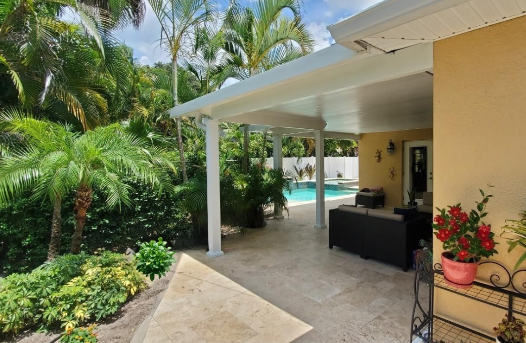 A backyard patio with a view of a swimming pool, surrounded by tropical plants and palm trees, with outdoor seating and decorative plants near the house.