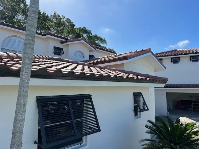 Photo of a white house with red tile roof, black window shutters, and a palm tree in the front yard, under a blue sky.