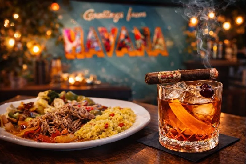 A plate of Cuban food with shredded meat, vegetables, and yellow rice, along with a cocktail garnished with a cherry and a cigar, in a Havana-themed restaurant with warm lighting and a blue wall with "Greetings from Havana" written on it.