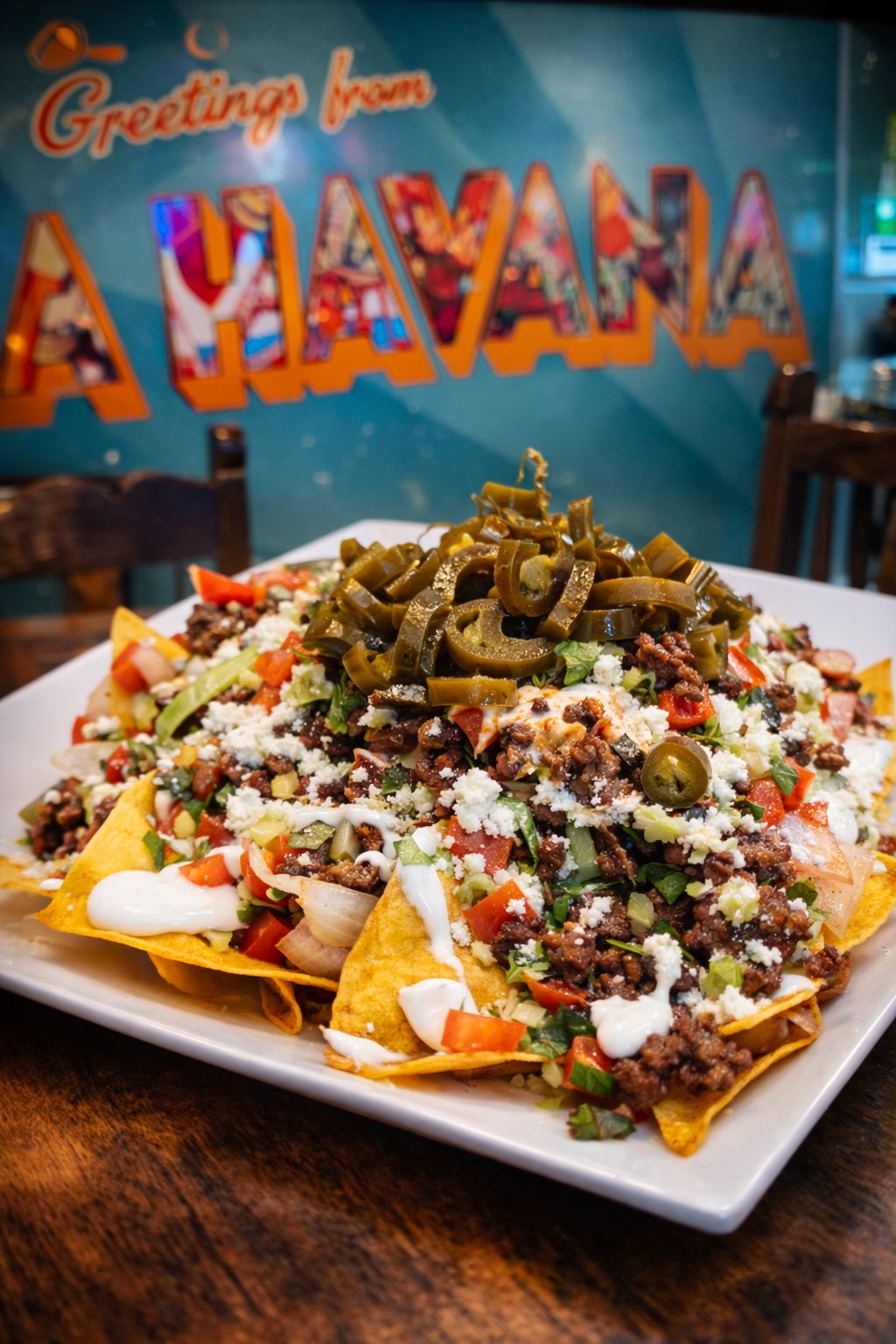 A plate of nachos topped with ground beef, shredded cheese, diced tomatoes, chopped green onions, sour cream, and sliced jalapeños, with a colorful 'Greetings from El Salvador' sign in the background.