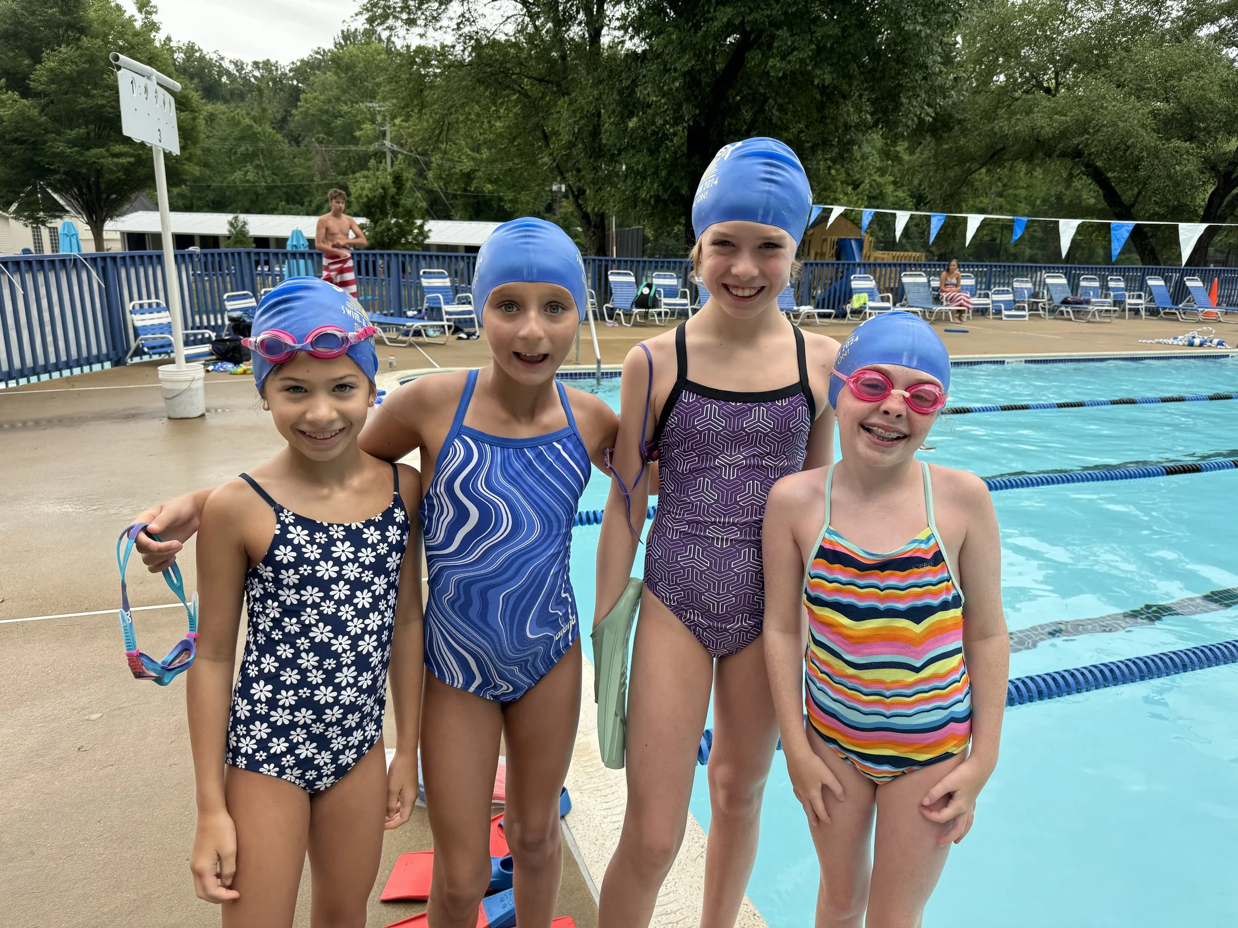 Four young girls in swimsuits and blue swim caps standing at the edge of a swimming pool, smiling for the camera. A boy in striped swim trunks is in the background.