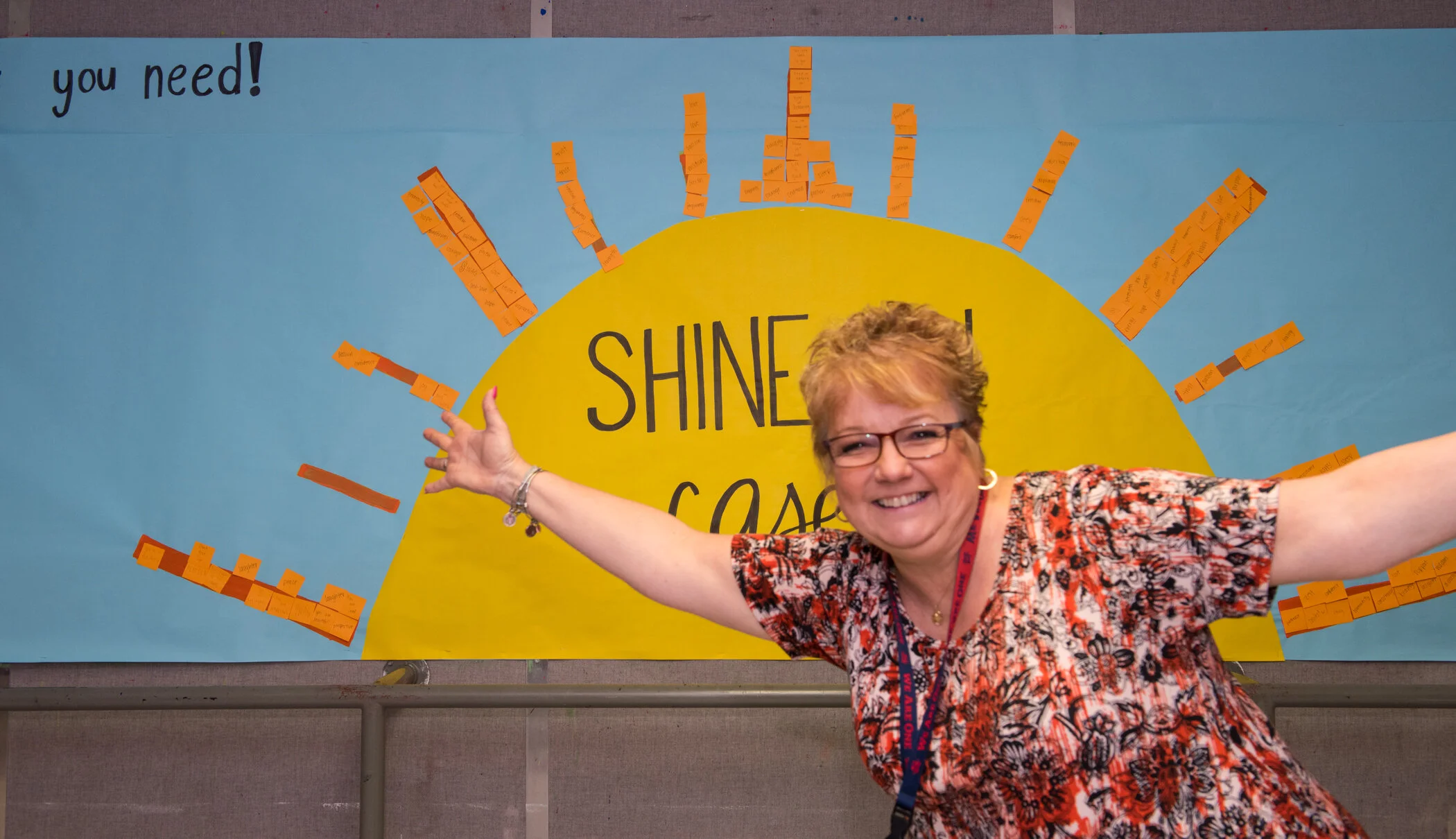 A smiling woman with glasses and a floral shirt, extending her arms in front of a blue poster with a yellow sun and the words 'SHINE ON!' in the center, surrounded by orange sticky notes.
