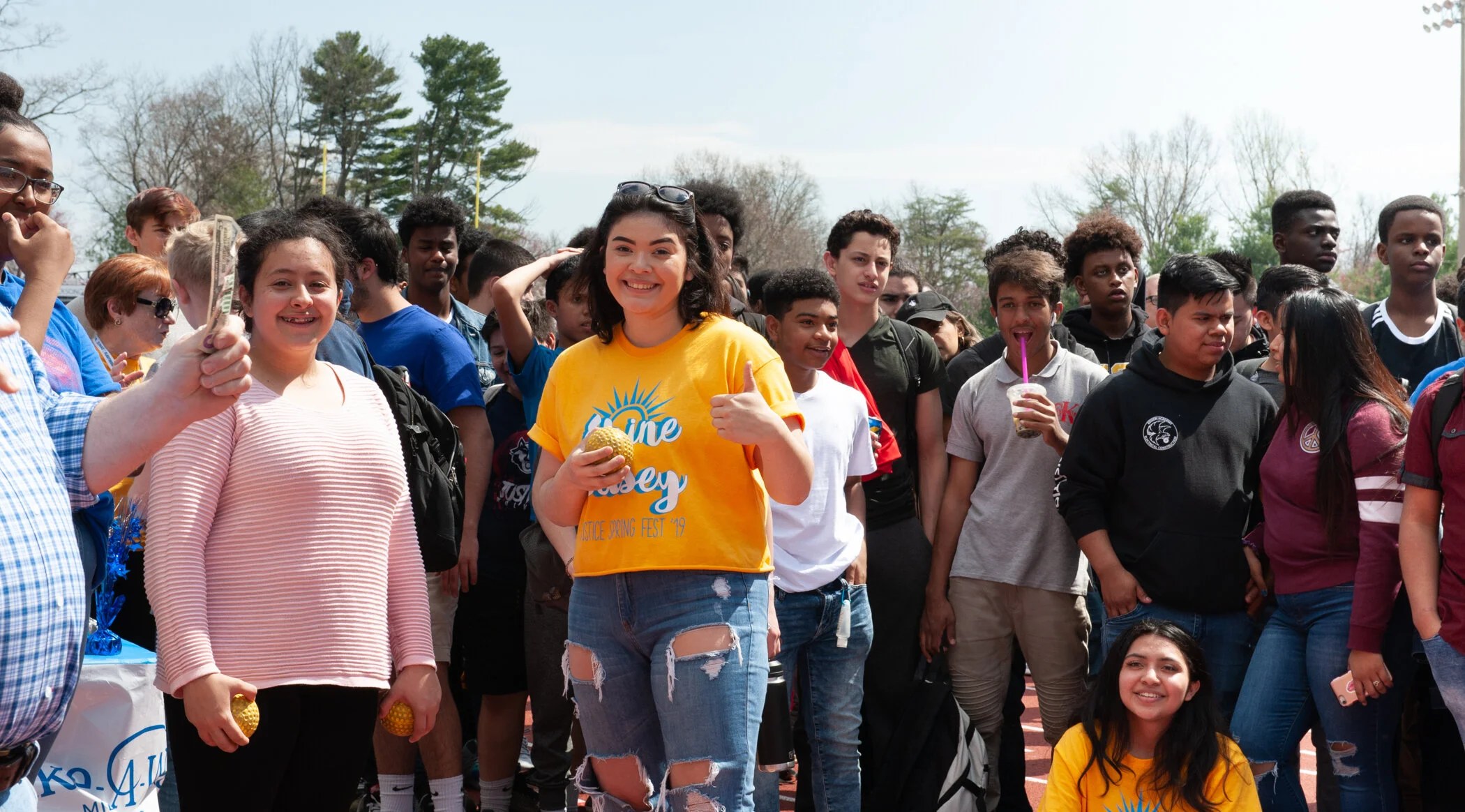 A group of smiling teenagers and young adults gathered outdoors on a cloudy day, some holding objects like pineapple-shaped items and drinks, participating in an event or gathering.