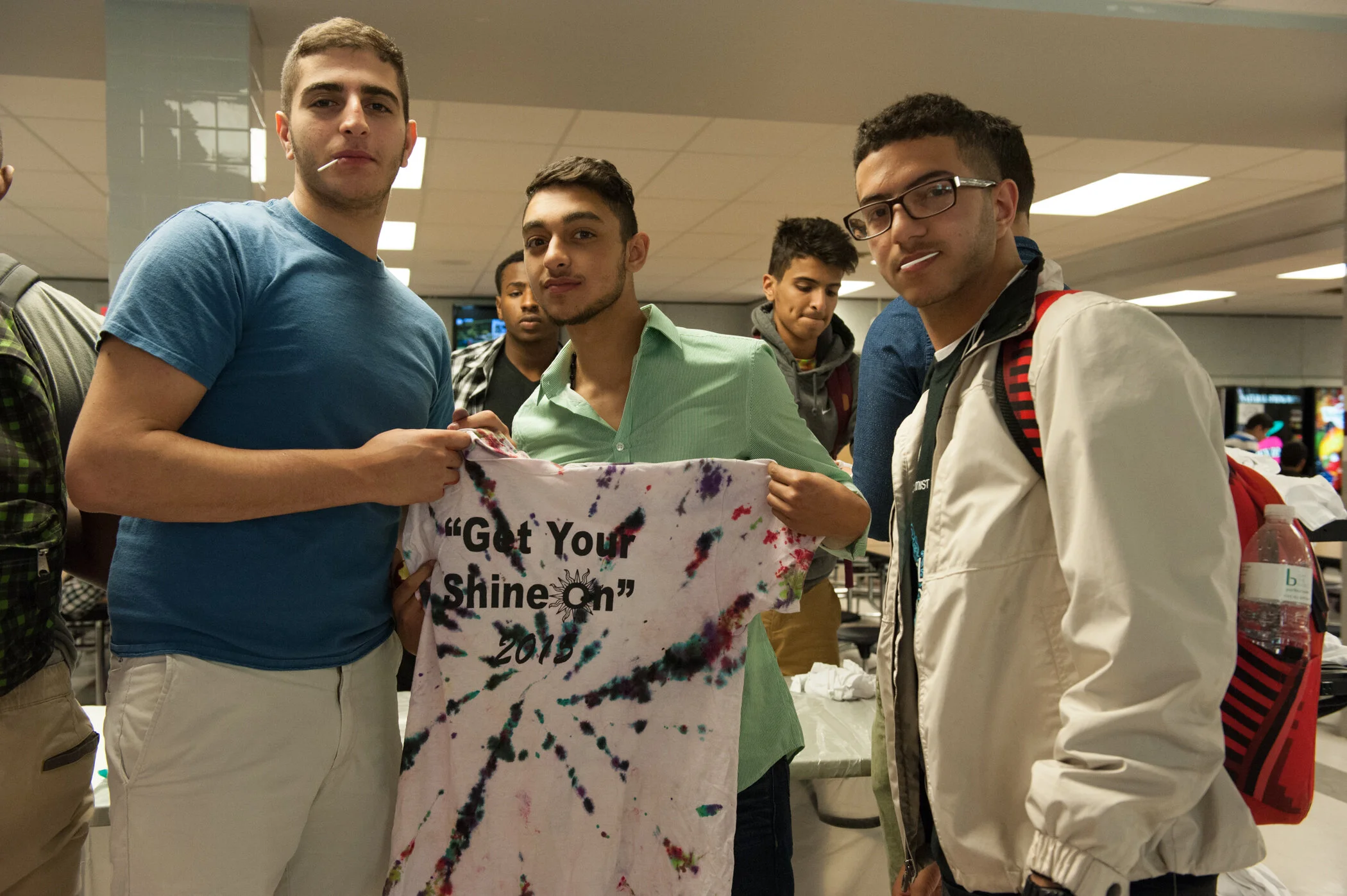 Group of young men at an indoor event, holding a tie-dye t-shirt with the text "Get Your Shine On" and the year "2018" printed on it.