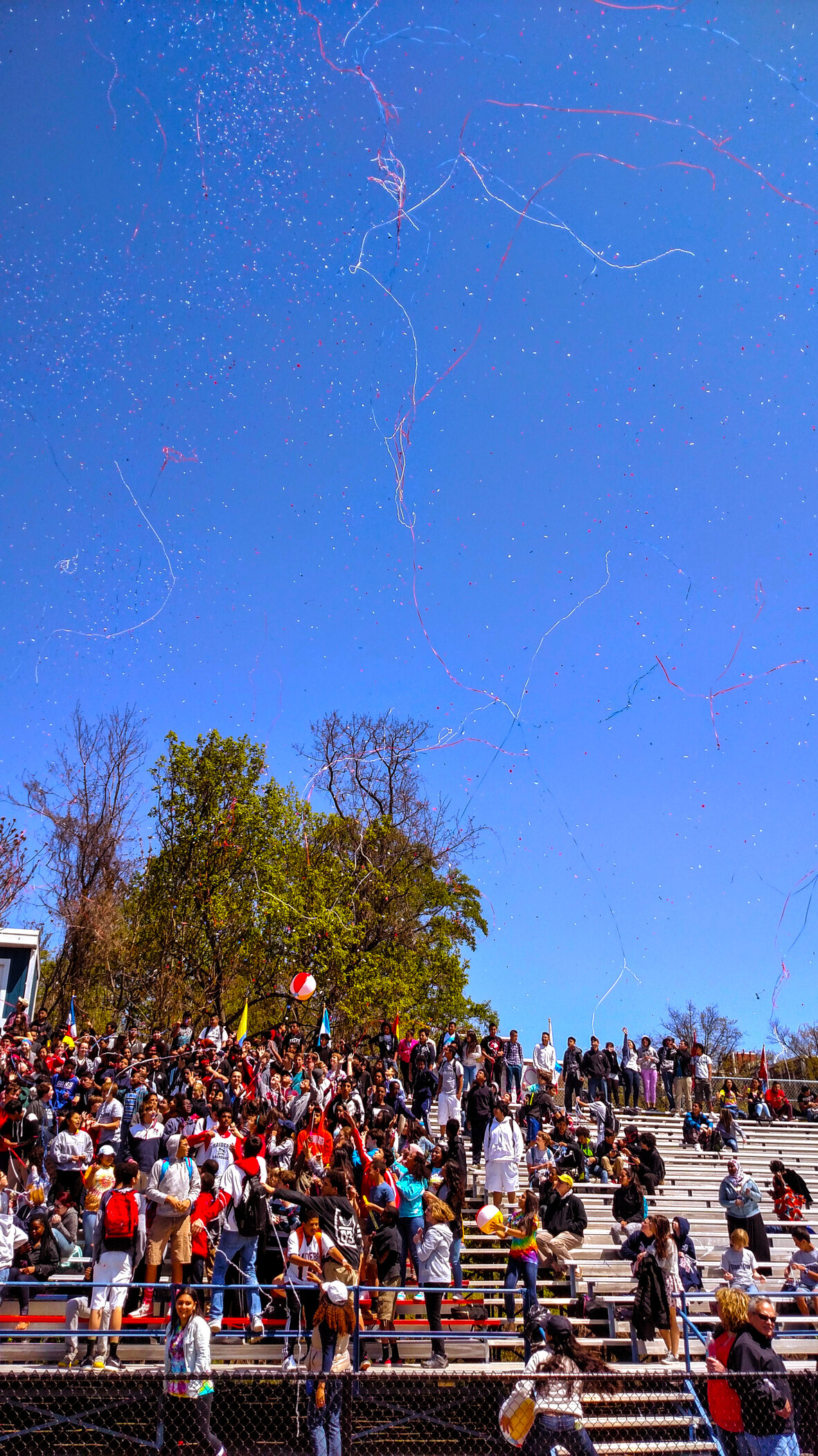 A large crowd of people gathered on tiered bleachers outdoors, releasing balloons into a bright blue sky with streaks of pink and blue streamers or ribbons.