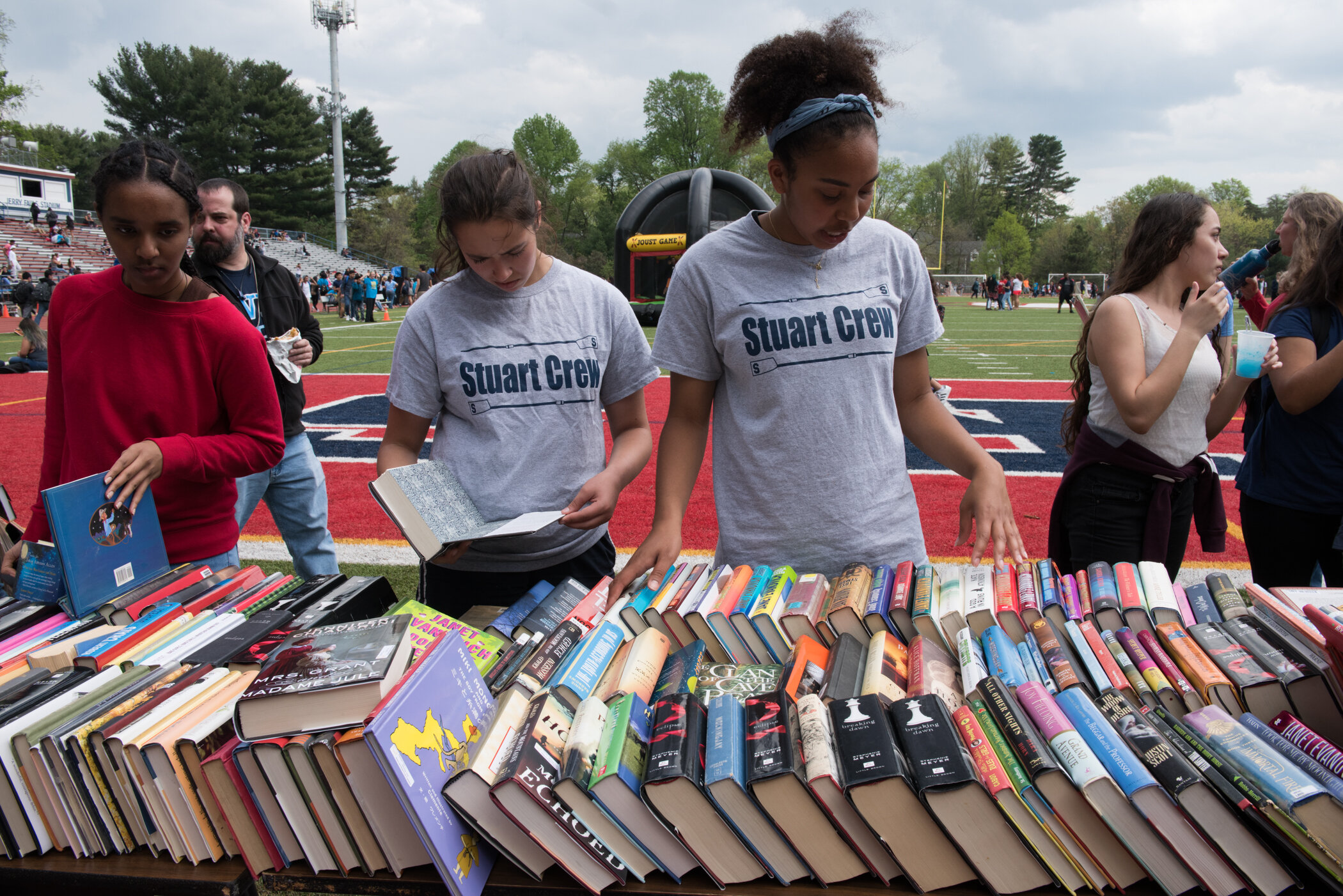 People browsing books at a book sale on a football field, with trees and spectators in the background.