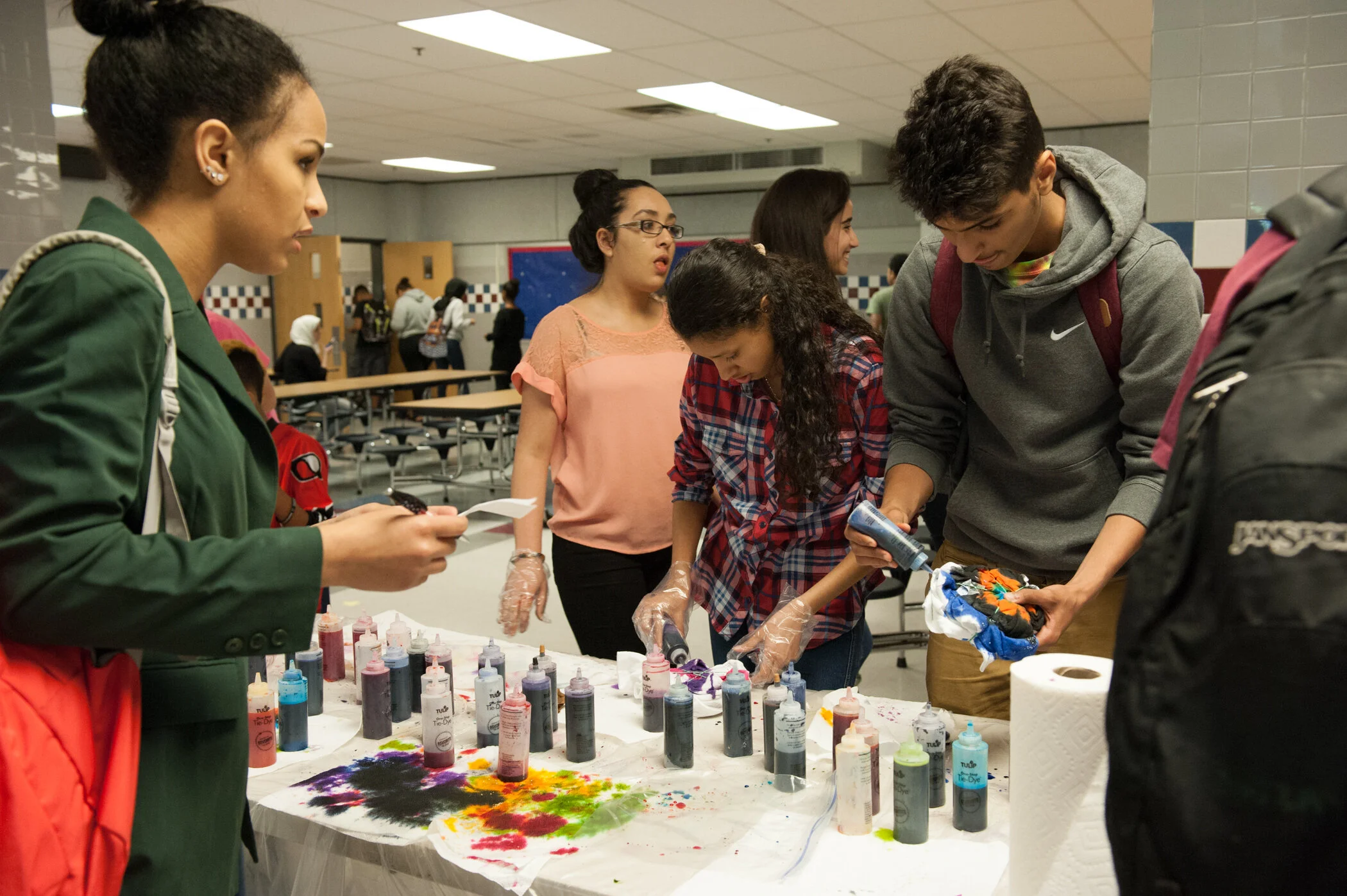 People gathered around a table with colorful ink bottles and paper towels, creating tie-dye or colorful fabric designs in an indoor setting.