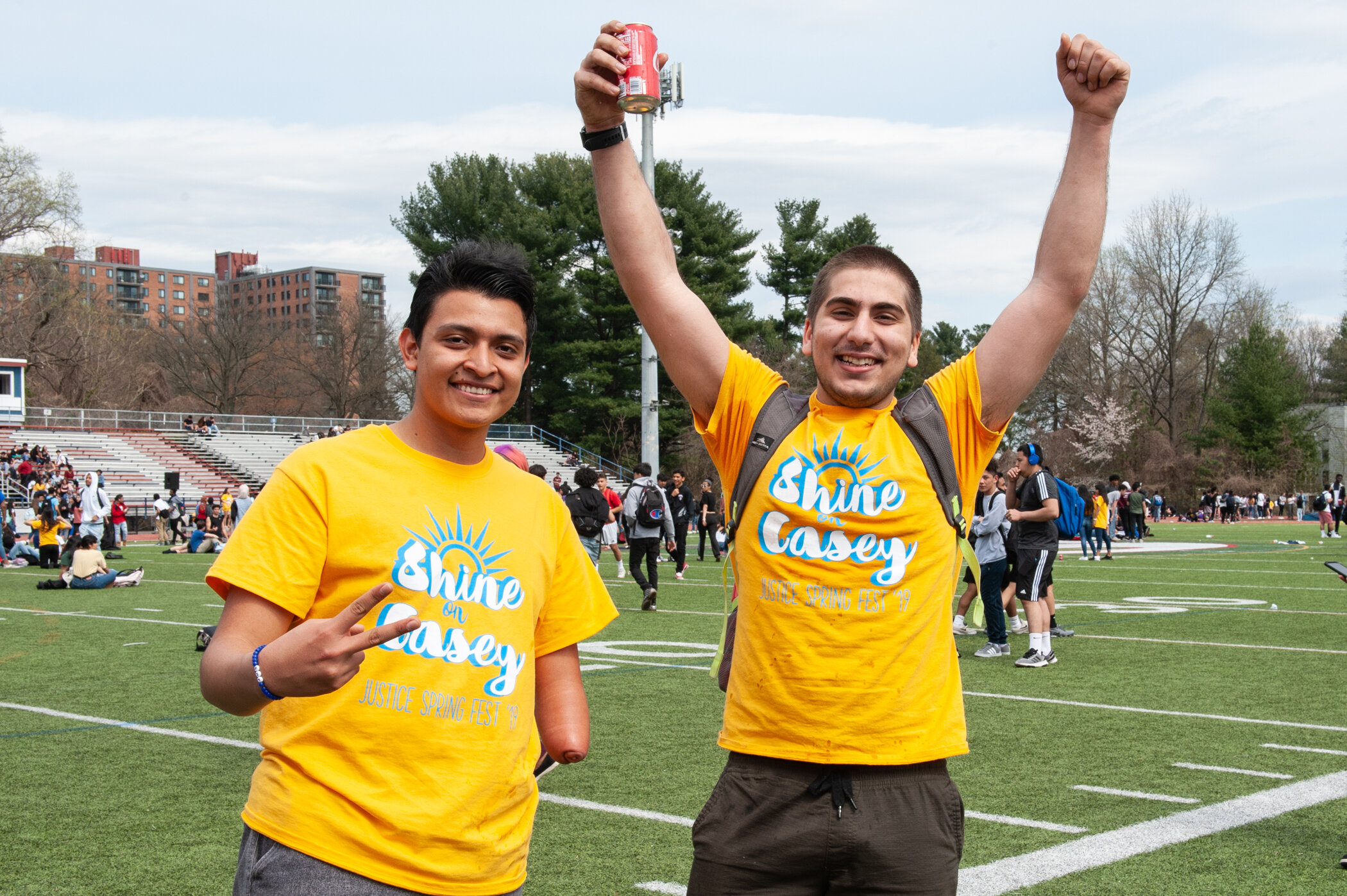 Two young men wearing yellow t-shirts with 'Shine on Casey' printed on them, standing on a football field during a spring fest, smiling, with trees and a crowd in the background.