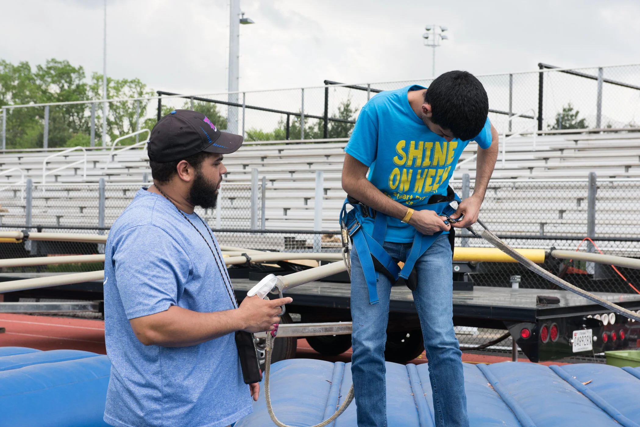 Two young men preparing for a bungee jump at an outdoor sports facility. One man is adjusting the harness on the other man, who is standing on an inflatable platform. The man standing on the platform is wearing a blue t-shirt that says 'SHINE ON WEB'
