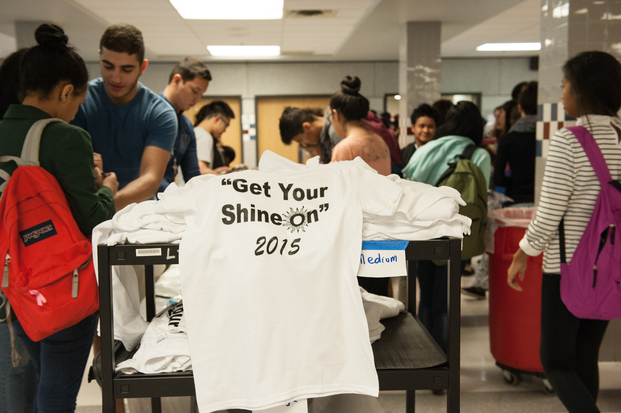 People in a busy indoor registration or event area, with a table of white T-shirts that say 'Get Your Shine On 2015' in black text.
