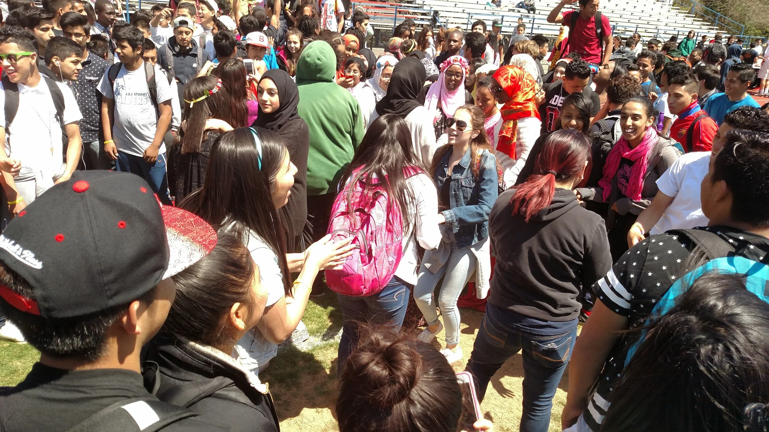 Crowd of diverse young students gathered outdoors, some with backpacks, engaging in a social activity on a sunny day, with bleachers in the background.