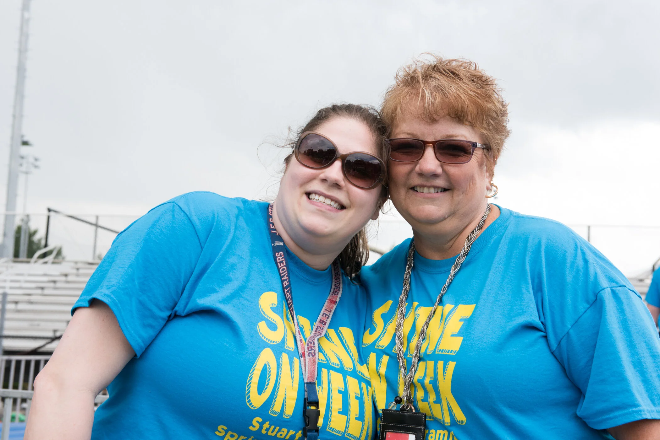 Two women smiling, wearing matching blue T-shirts and sunglasses, standing outdoors with bleacher seats in the background.