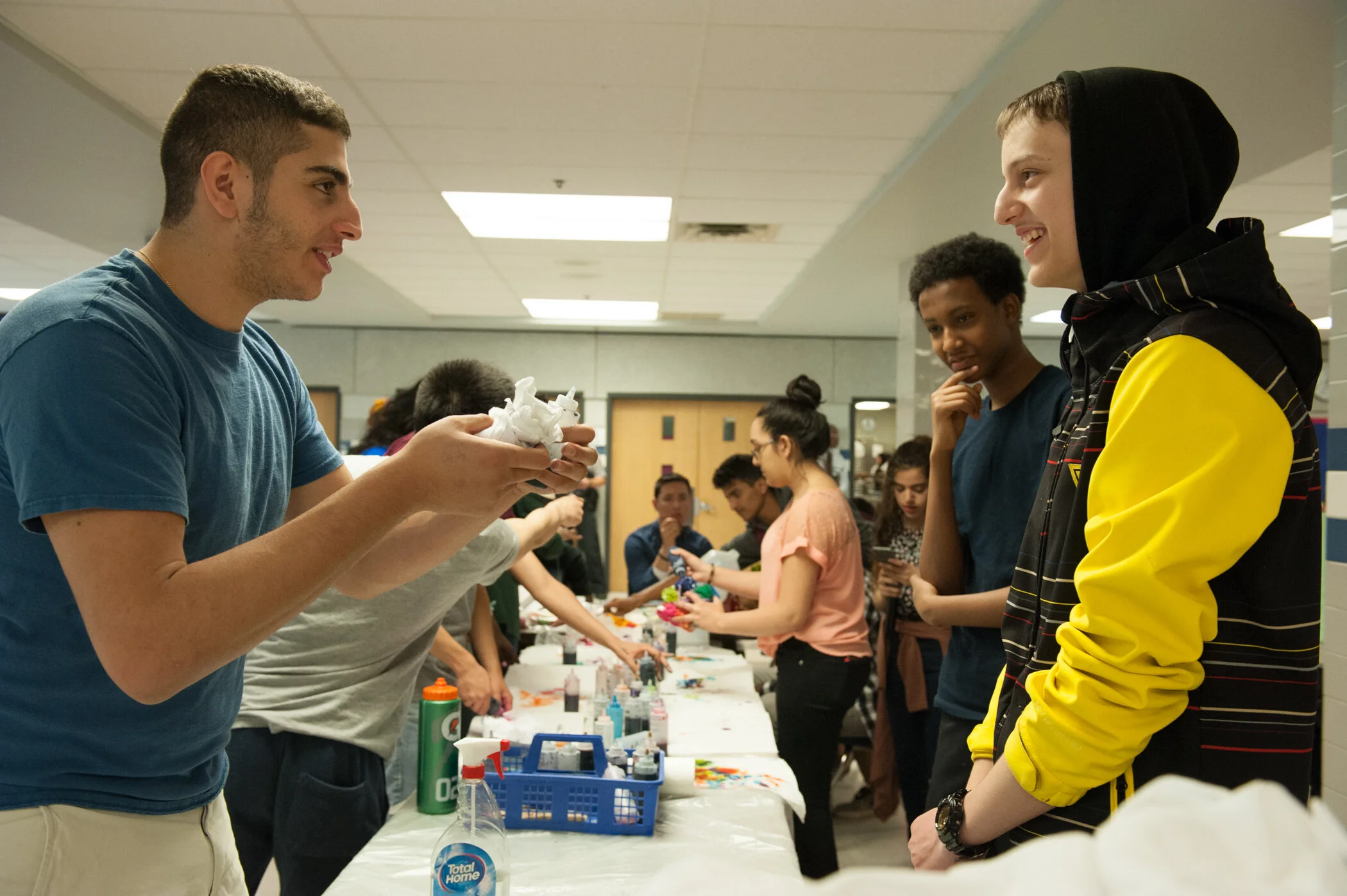 Young man at a table demonstrating arts and crafts to a teenager with a striped jacket inside a school hallway.