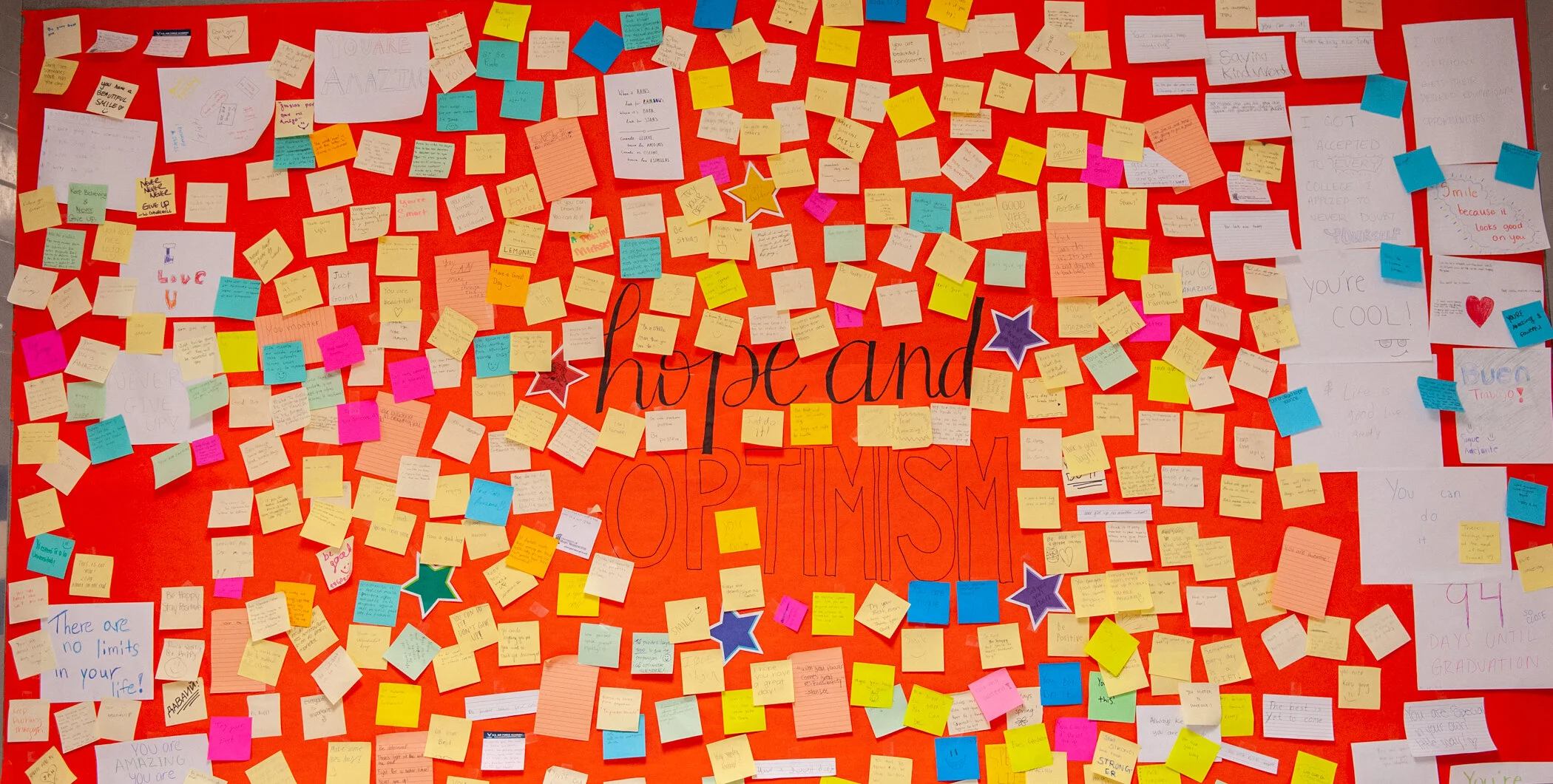 A vibrant bulletin board filled with numerous colorful sticky notes and handwritten messages of encouragement and positivity, with a central large sign that reads 'hope and OPTIMISM' on a red background.