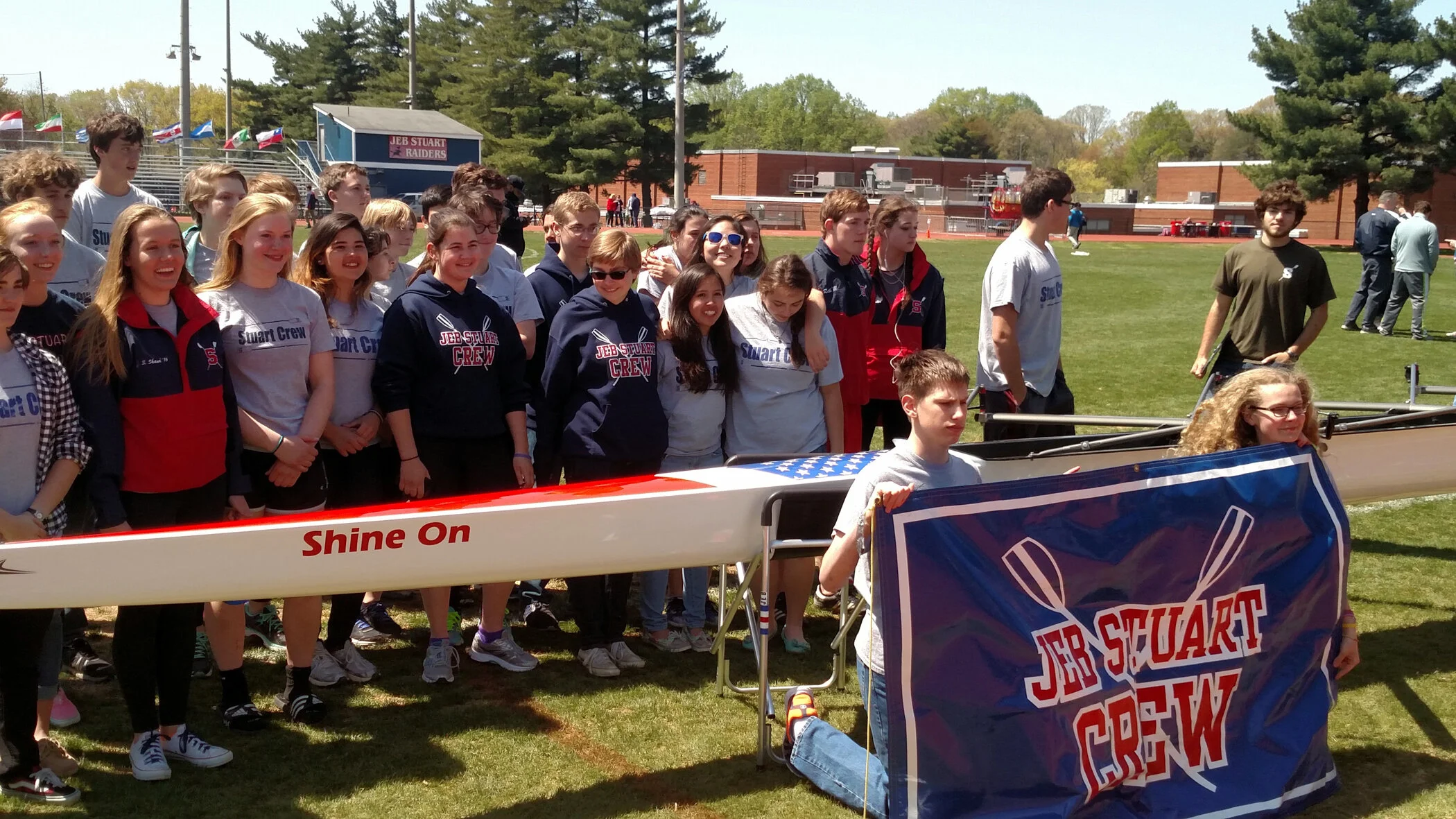 A group of students and coaches gathered on a grassy field, with some holding a blue banner that reads 'JER STUART CREW' and a boat labeled 'Shine On' in front of them, at a school or community sporting event on a sunny day.
