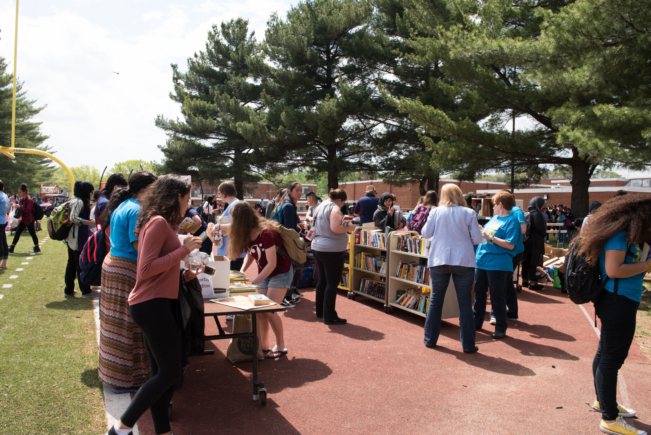 A group of people browsing books at an outdoor book sale under large trees on a school track field.